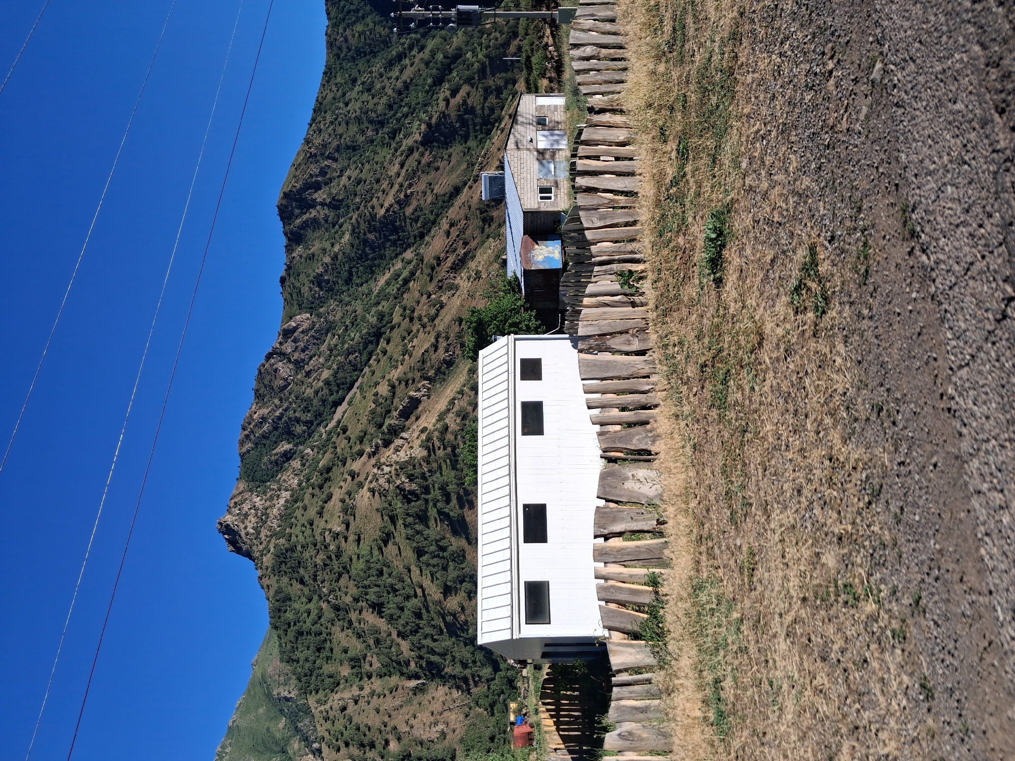The image, taken in Shatin, Armenia, depicts a mountainside scene under a clear, bright blue sky, suggesting daytime and fair weather. The primary subjects are two small buildings situated on a steep, verdant mountain slope. One building is a modern, white, rectangular structure with three black rectangular windows. Adjacent to it is an older, multi-colored building with a blue-tiled roof and several windows, appearing to be in a state of disrepair or undergoing renovation.
A pathway made of rough-hewn wooden logs ascends the slope alongside the buildings, bordered by dry, golden grass and loose gravel. In the background, the rugged mountainside is covered in dense green foliage and rocky outcrops. Power lines stretch diagonally across the upper left portion of the image, a common sight in mountainous regions.
There is no visible text in the image. The scene conveys a sense of remote, rugged beauty and a functional, perhaps slightly rustic, human presence in a natural environment. The presence of the buildings suggests habitation or a point of interest within the mountainous landscape. The lighting indicates it is likely mid-morning or afternoon.