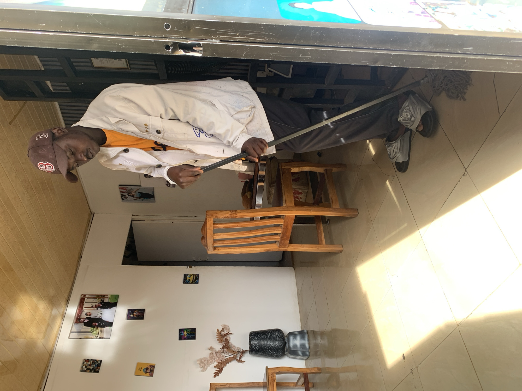 The image displays an indoor scene, likely a shop or small business in Jos, Nigeria, featuring a man engaged in an activity. The man, with dark skin, is wearing a brown baseball cap with white Boston Red Sox "B" and "SOX" logos. He has an orange t-shirt under a light-colored, possibly white or off-white, jacket or work coat featuring three stars embroidered on the right chest. He wears dark trousers and grey or silver slides.
He stands beneath a dark, metallic structure, possibly a counter or a part of a machine, holding a long, thin metallic tool or measuring stick with both hands. His gaze is directed downwards, suggesting concentration on his task. Near his feet, a mop head is visible on the floor.
The setting includes white walls, with wooden paneling on the left side of the room. The floor is tiled with light-colored, possibly cream, square tiles arranged diagonally. A bright patch of sunlight illuminates the floor, indicating it is daytime. The walls are decorated with several framed pictures or prints, including a larger one depicting a group of people, and smaller ones featuring abstract designs, a smiling face, a "FREEDOM" text, a "BATMAN" logo, and "DAB" text. On the floor, two decorative planters or vases, one dark speckled and one grey patterned, hold artificial branches with light-colored flowers or leaves. A light brown wooden chair or stool is also present.