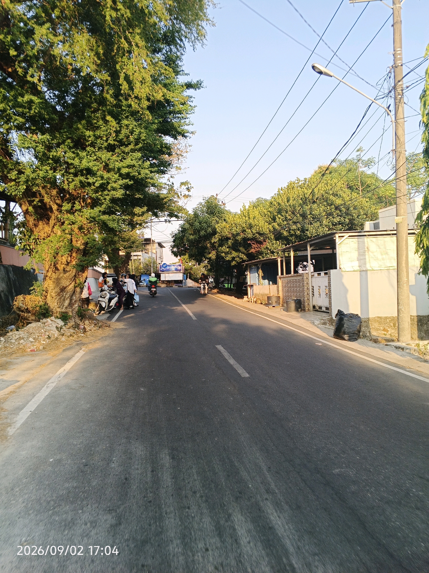 The image depicts a street scene in Pare-Pare, Indonesia, on September 2, 2026, at 17:04. A paved road runs through the frame, bordered by trees and buildings on either side. On the left, a large tree with dense green foliage stands prominently near the roadside. A group of people are gathered around several motorcycles parked under the tree. Further down the road, more individuals and motorcycles are visible, suggesting a moderate level of activity. On the right side of the road, a building with a light-colored facade and a covered outdoor area is present. A large black trash bag is placed near the sidewalk in front of this building. Utility poles with numerous wires are also visible, indicating the presence of infrastructure. The sky is clear and blue, with the sun casting warm light on the scene, suggesting it is late afternoon. The overall impression is of a typical urban street environment in Indonesia.
