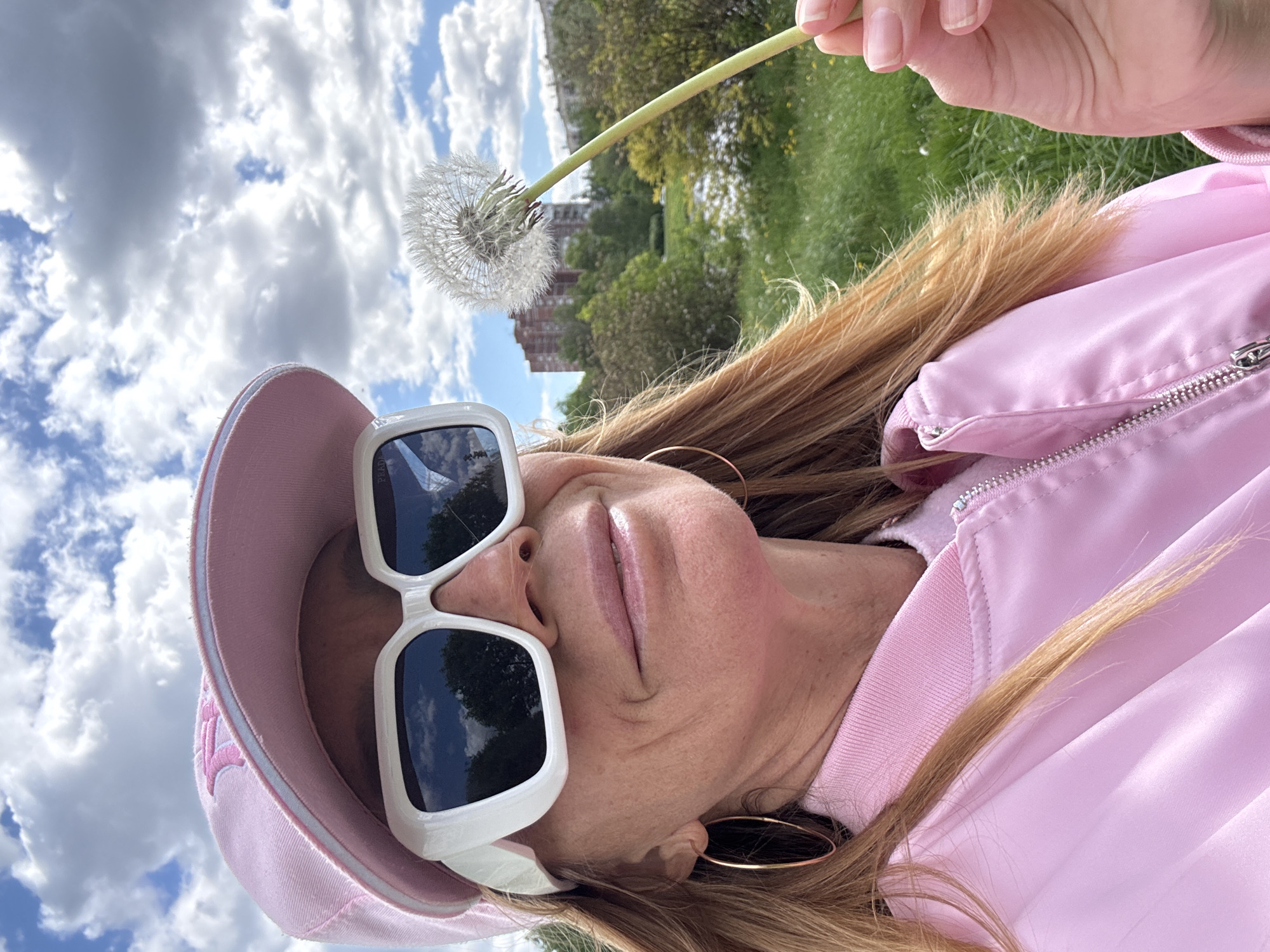 A person, possibly female, wearing a pink cap, white sunglasses, and a pink jacket, is holding a dandelion against a backdrop of sky with clouds and greenery. The photo appears to be taken outdoors in a park or garden setting. Based on visible buildings in the distance, it is possible the location is Saint Petersburg, Russia, but this is difficult to confirm definitively without further context.