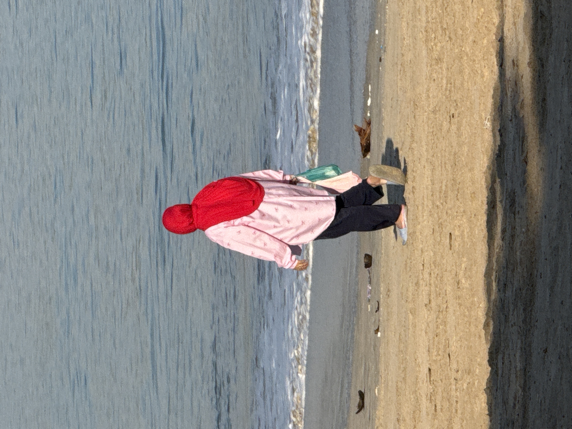 A person wearing a bright red hijab and a pink patterned long-sleeved shirt walks along the sandy shore of a beach. Dark blue pants are visible below the shirt. The person carries a green plastic bag in their right hand and a pink bag in their left. They are walking away from the camera towards the water. Gentle waves are lapping the shore. The sand is tan and appears dry. Several small, dark objects, possibly shells or debris, are scattered on the sand. The water is a muted blue-grey, with a line of white foam indicating the breaking waves. The foreground of the image is in shadow, cast from an unseen object to the left, obscuring the lower portion of the sand and the person's feet. The overall scene is one of a solitary walk on a beach, likely during daylight hours.