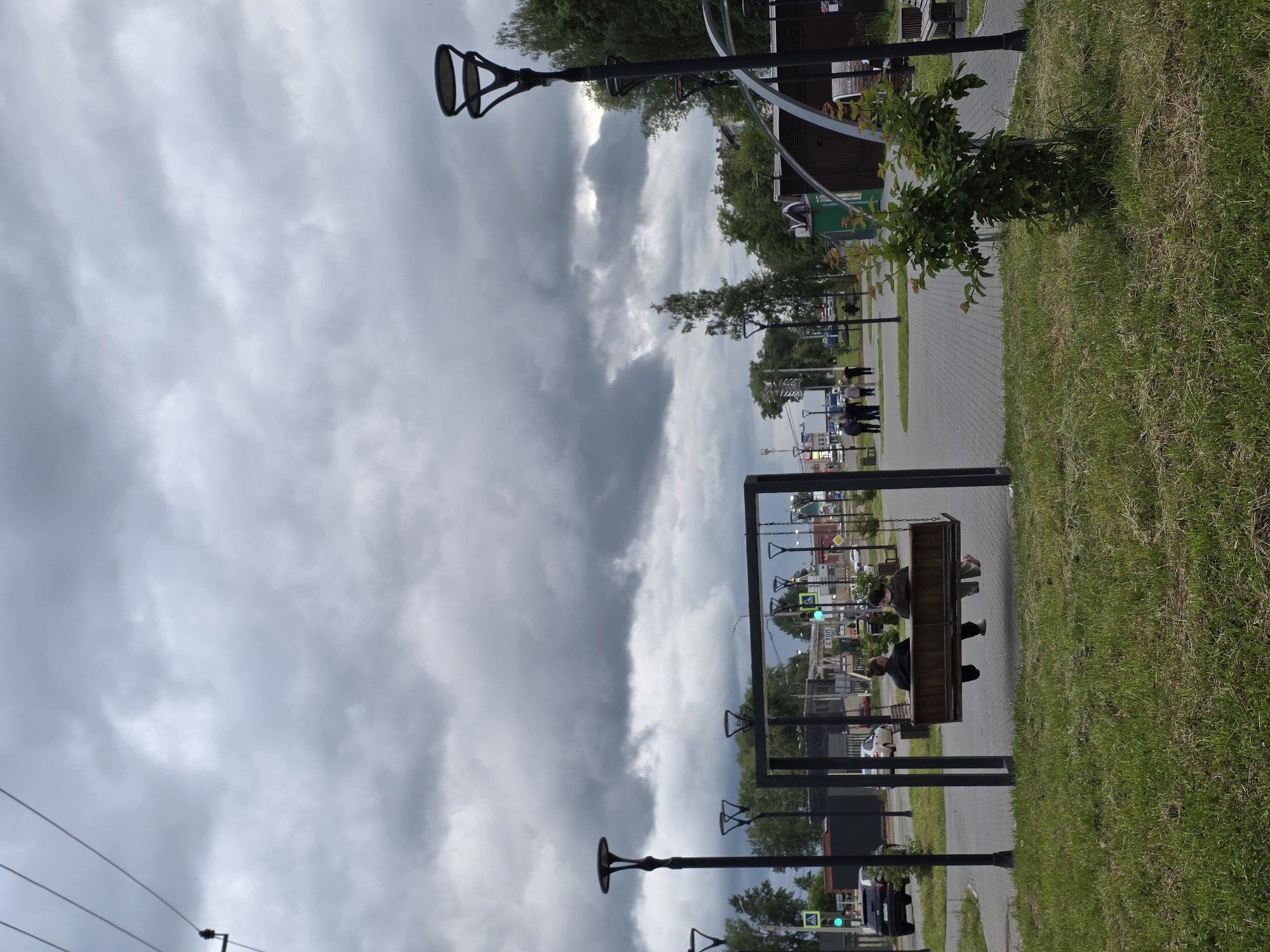 This image depicts a public square or park area in the city of Chaikovsky, Russia, under an overcast sky dominated by heavy, dark grey clouds. The foreground features green grass, with a small, leafy bush on the right. In the midground, a dark-framed wooden swing set is positioned on a paved area made of interlocking pavers. Two individuals are seated on the swing, facing away from the camera. The person on the left wears dark clothing and has dark hair tied up, while the person on the right also wears dark clothing and has lighter hair.
Multiple modern, black street lamps with distinctive two-tiered, disc-like tops are scattered throughout the scene. Beyond the paved area, a road is visible with a black SUV on the left and a white sedan further back. Traffic lights displaying green signals and pedestrian crossing signs are present. Various buildings, including commercial structures and residential properties, line the street. A sign with the Cyrillic word "СТОП" (STOP) is discernible near the road. Several people are walking on a paved path in the background. Green trees and other vegetation are present behind the buildings and along the edges of the park. Power lines are visible in the upper left corner of the frame.