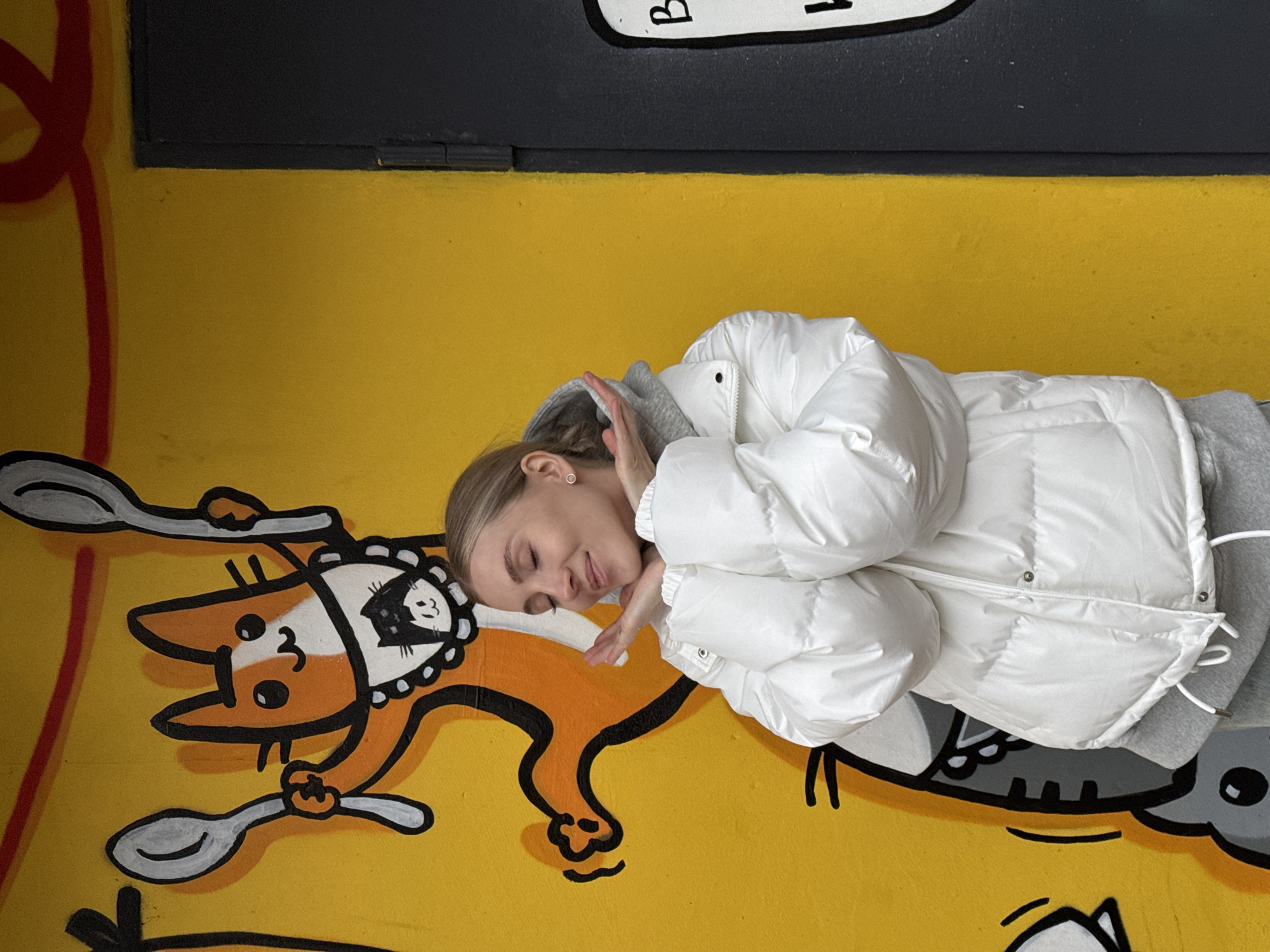 A woman in a white puffer jacket is lying next to a mural on a yellow wall. The mural features an orange cartoon cat holding two spoons. This mural is located in Izhevsk, Russia. The woman has her hands raised near her face and is smiling. She is also wearing a gray hooded sweatshirt. The wall is yellow with some graffiti markings.