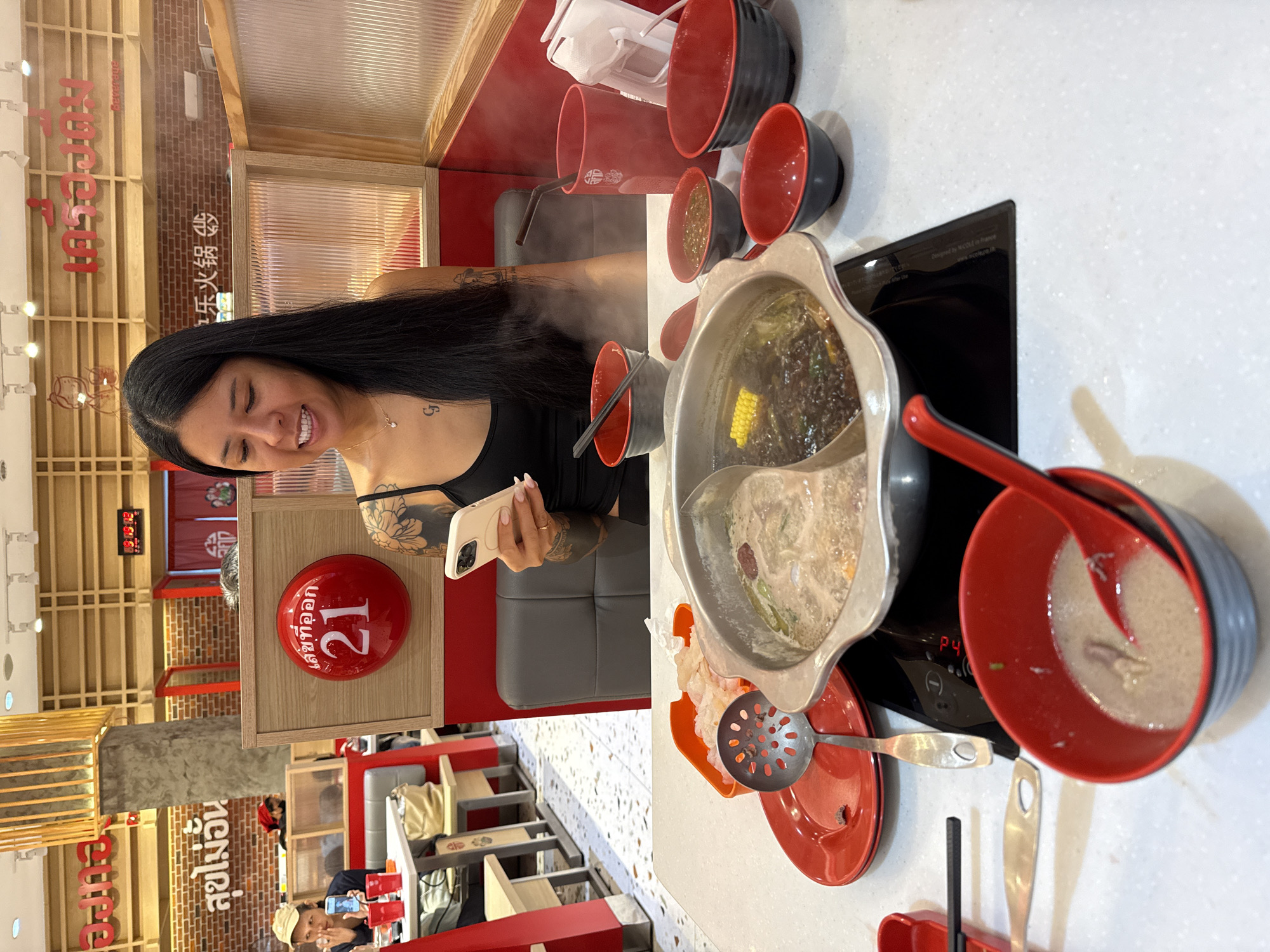 A woman with long, dark hair smiles and holds a phone in her left hand while seated at a restaurant table with a hot pot. The hot pot, which has two compartments, is on a black electric stove. Bowls, ladles, and a skimmer surround the pot. The restaurant has a red and tan color scheme. Red spheres with white Thai writing that reads “เลขที่ออก 21” are seen on the wall. The words “樂火鍋 泰国曼谷สุขไม่อั้น" are also visible. The location appears to be Bangkok, Thailand.