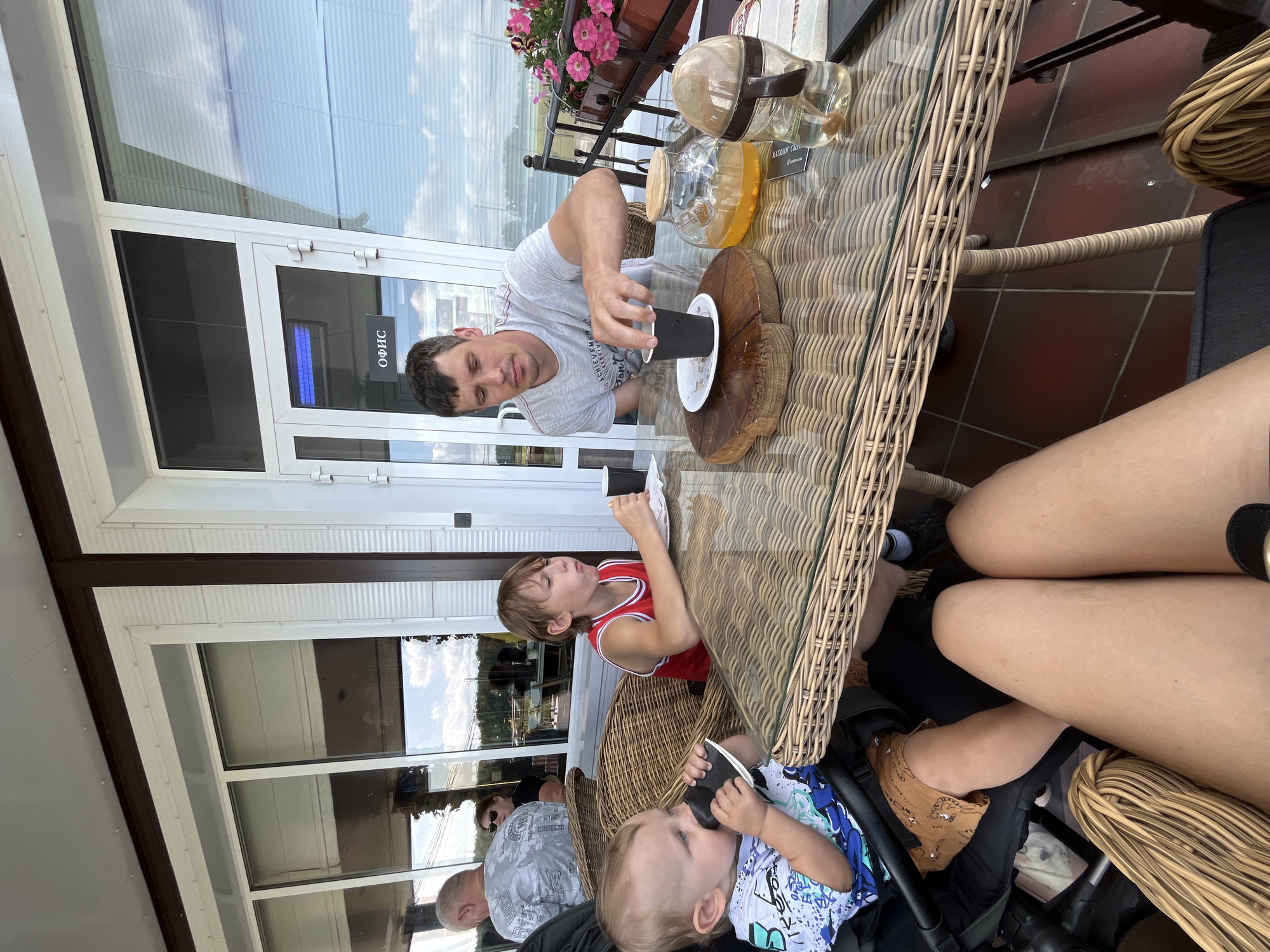 The image depicts an outdoor seating area at a cafe, likely in Akhlebaikha (Kovriginskii S/S), Russia. A man in a grey t-shirt is seated at a wicker table, reaching for a black cup. On the table, there is a wooden serving board, a glass teapot with orange liquid, and a baby bottle. A young boy in a red and white tank top is also seated at the table, eating. A baby in a stroller is to the left of the frame, holding a black cup to its mouth. In the background, large windows with blinds suggest an indoor space, and a sign on the door reads "ОФИС" (Office). The overall environment appears to be a casual dining setting, possibly by a body of water, given the hazy landscape visible through the windows.