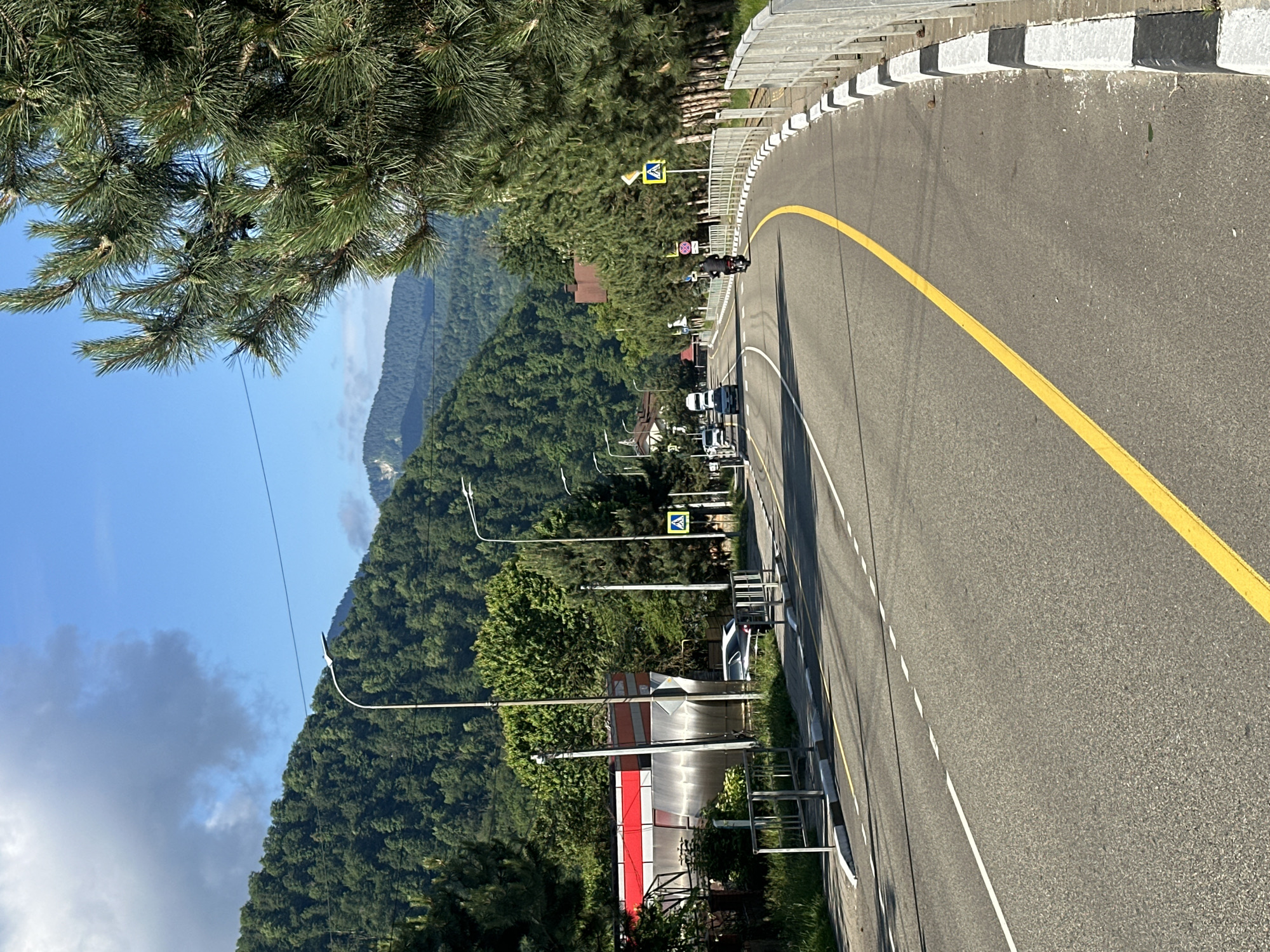 The image depicts a street scene in Krasnaya Polyana, Russia, on a sunny day with some clouds. A two-lane asphalt road curves to the right, marked with a solid yellow line on the right side and dashed white lines separating the lanes. On the left side of the road, there's a building with a red and white facade, a bus stop shelter with metal railings, and a parked car. Further down the road, a few more vehicles are visible, including a white minivan and a dark-colored car. A motorcyclist wearing a black jacket is riding on the right side of the road. Pedestrian crossing signs with yellow diamond shapes and black symbols are posted on the right side of the road. A metal fence with white and black striped concrete bollards lines the right edge of the road. The background is dominated by lush green mountains covered in dense trees, indicating a mountainous or hilly terrain. Tall pine trees are visible on the right side of the road. Streetlights are positioned along the road.