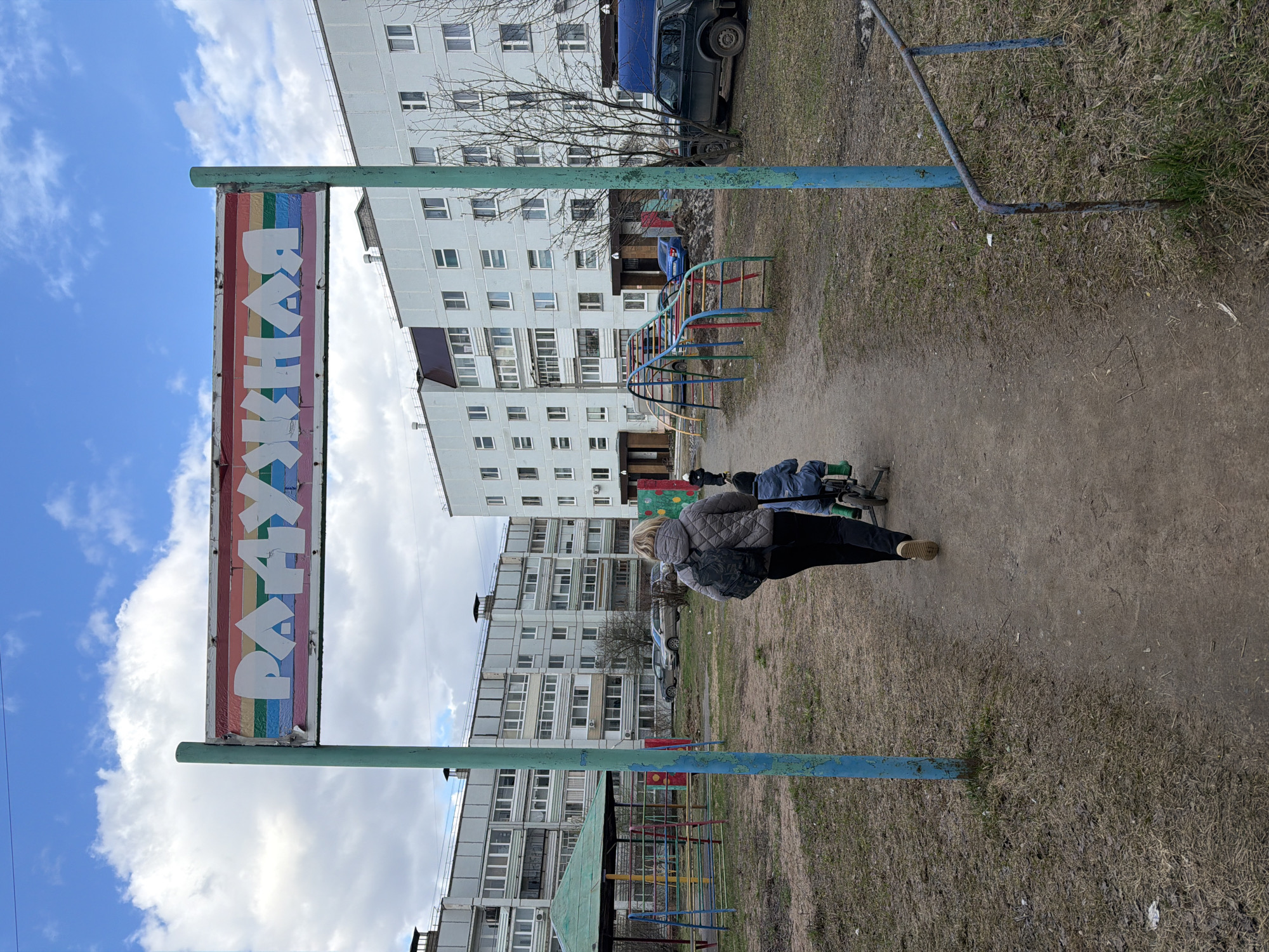 The photograph depicts an urban scene in what is likely Chaikovsky, Russia, based on the Cyrillic script "РАДУЖКА" on the sign, which translates to "Rainbow." The image is oriented vertically. In the foreground, there's a playground sign with rainbow stripes and Cyrillic lettering. The sign is supported by blue metal poles.
A woman is walking away from the camera, pushing a baby carriage on a dirt and grass surface. She is wearing a gray quilted jacket, black pants, and tan shoes, and has a backpack.
Behind the woman and playground area is a multi-story apartment building with numerous windows. A few vehicles are visible near the building. The sky is partially cloudy. The general environment suggests a residential area during the daytime. The playground is somewhat worn, indicating regular use.