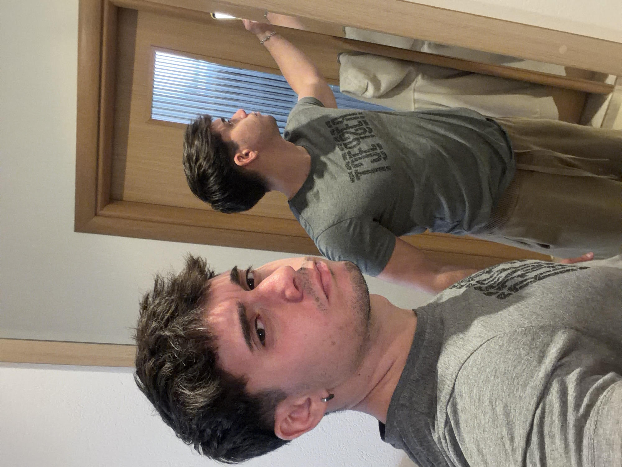 A man is taking a selfie in a mirror. He is wearing a grey t-shirt. The mirror is mounted in a wooden frame. The reflection shows a second person wearing a grey t-shirt and khaki pants. The reflection also shows a window with horizontal blinds and what appears to be a pillow on a bed or couch. There is no evidence to suggest that this image was taken in Blagoevgrad, Bulgaria.