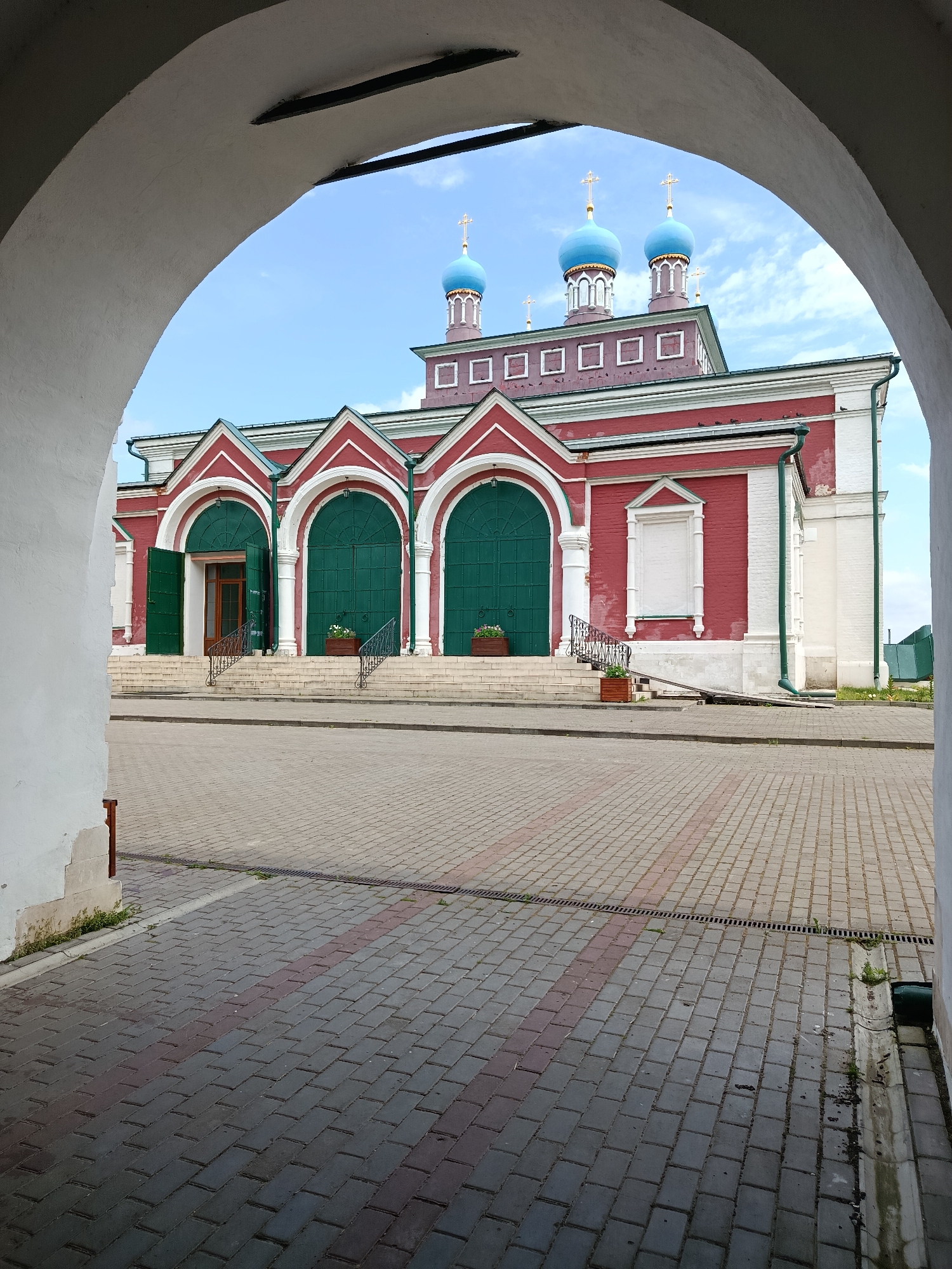 The image captures a view through a large, white arched entryway towards a red brick building with green doors and blue domes, likely a church or monastery, in Radovitsy, Russia. The building features multiple arched entrances with large, dark green double doors, some of which are open. White columns flank these entrances, and a set of stone stairs leads up to them. Above the red brick facade, a section of the building is topped with a darker colored roof, and on top of that, several blue domes with golden crosses rise against a bright blue sky with scattered white clouds. The ground in front of the building is paved with gray and reddish brickwork in a patterned layout. A drain grate runs along the edge of the paved area. The foreground is dominated by the interior of the white arched structure, with a hint of a wooden post visible on the left. The overall scene depicts an architectural site bathed in daylight.