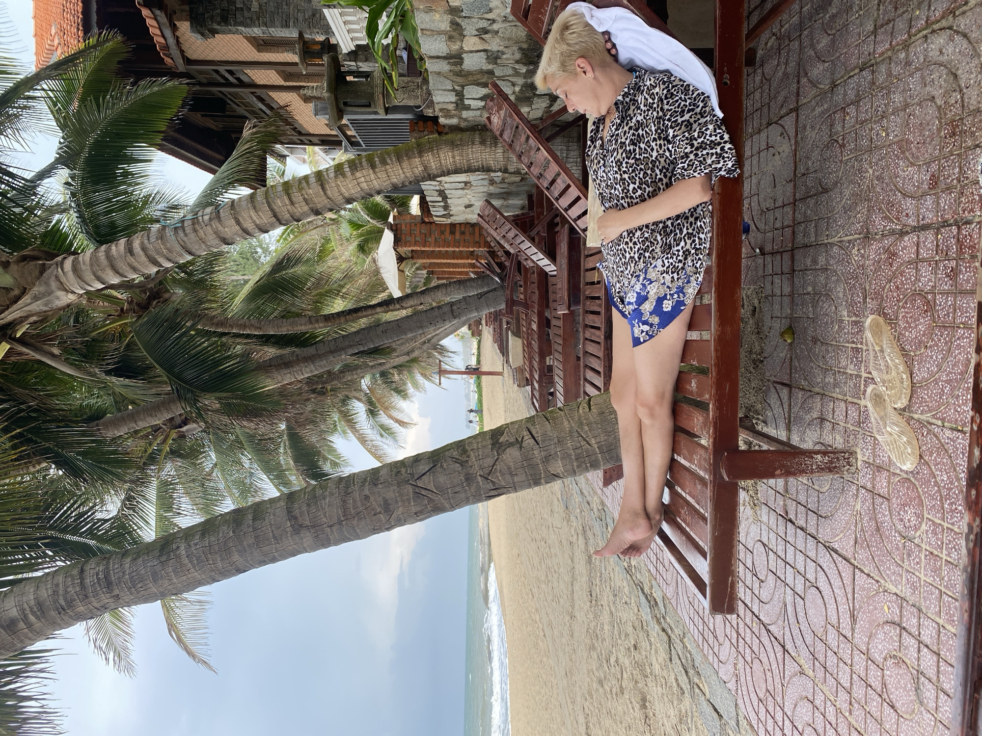 The image depicts a serene tropical beach resort scene, set in what appears to be Phu Thuy Ward, Vietnam. A fair-skinned woman with short blonde hair is seated on a wooden lounge chair, looking down in a relaxed posture. She is wearing a short-sleeved leopard print shirt with blue shorts that have a floral pattern. A white towel is draped over her head and resting on the back of the chair. Her bare feet are visible, resting on the chair's slats.
To the right, on the ground, a pair of clear or light-colored sandals are placed on a distinctively patterned red and brown tiled surface, which features a repeating circular motif. Several tall palm trees with rough, textured trunks and vibrant green fronds are prominent throughout the scene, suggesting a lush, tropical environment. In the background, the sandy beach stretches towards the ocean, where gentle waves are visible breaking near the shore under a partly cloudy sky. Further back, traditional-style buildings with red-tiled roofs and stone-clad walls are nestled among the trees, indicating a resort setting. The scene conveys a sense of peaceful relaxation during the daytime. No visible text is present in the image.