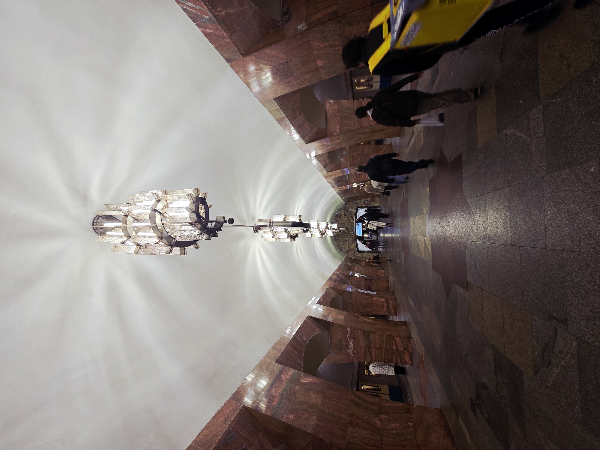 The image captures the interior of a subway station in an unknown city. The walls are lined with reddish-brown marble tiles, and the ceiling is white and vaulted. A row of elaborate chandeliers hang from the ceiling, providing illumination. Several people are walking through the station, with some wearing masks. The floor is also made of marble tiles in various shades. A blurred yellow object, possibly a taxi, is visible at the top right of the frame, adding a sense of urban activity.