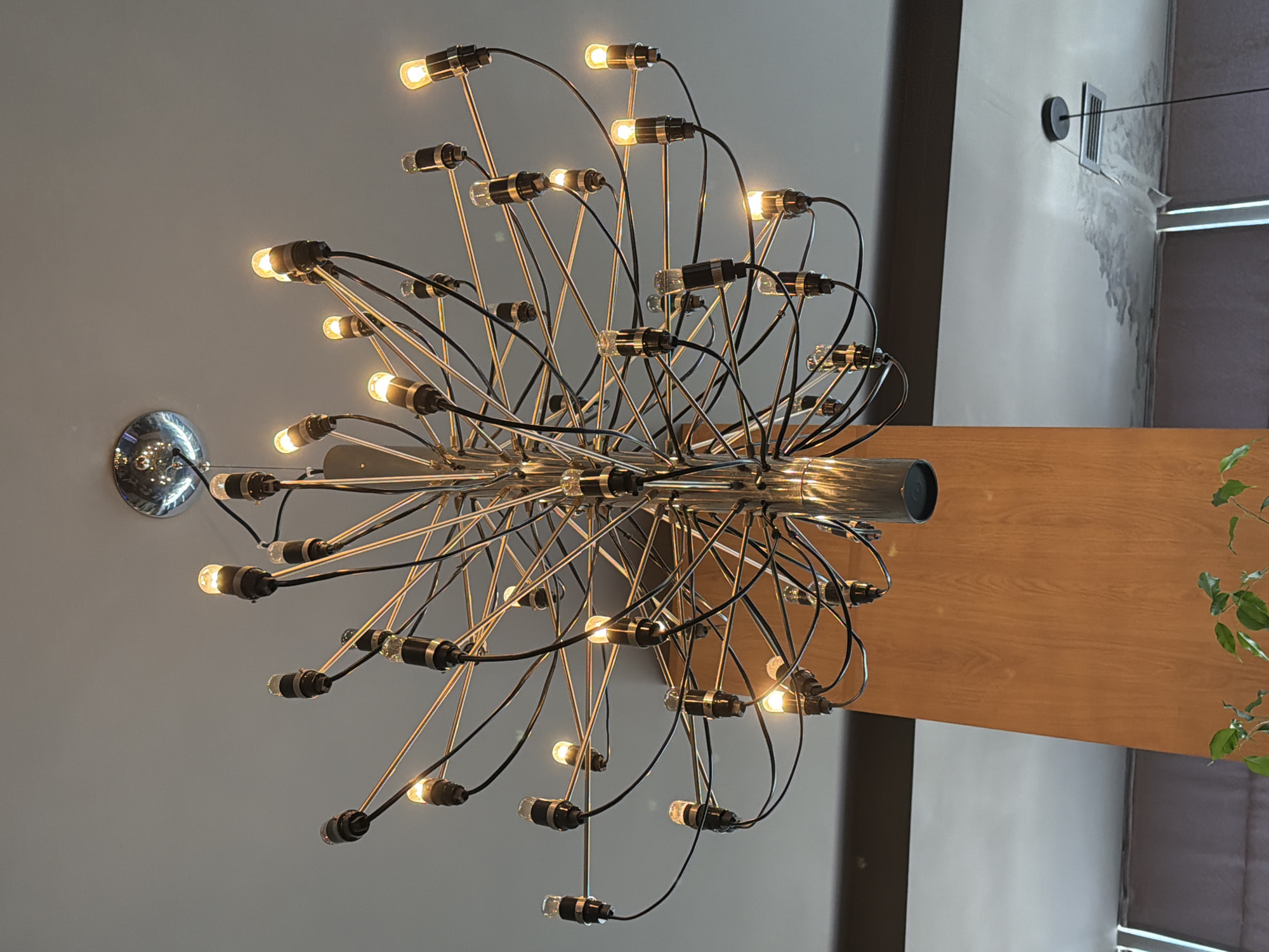 A modern, multi-tiered chandelier hangs from a textured white ceiling. The chandelier has a chrome finish and features numerous thin metal arms extending outwards, each holding a small, cylindrical light bulb. Black wires are neatly organized, connecting the bulbs to the central fixture. The chandelier is the primary focus, with its intricate design and warm, glowing lights. In the background, a light wood panel covers a structural column, and a section of a grey ceiling is visible with a vent and a small black hanging light fixture. The overall impression is one of contemporary interior design in an indoor space, likely a restaurant or hotel lobby.