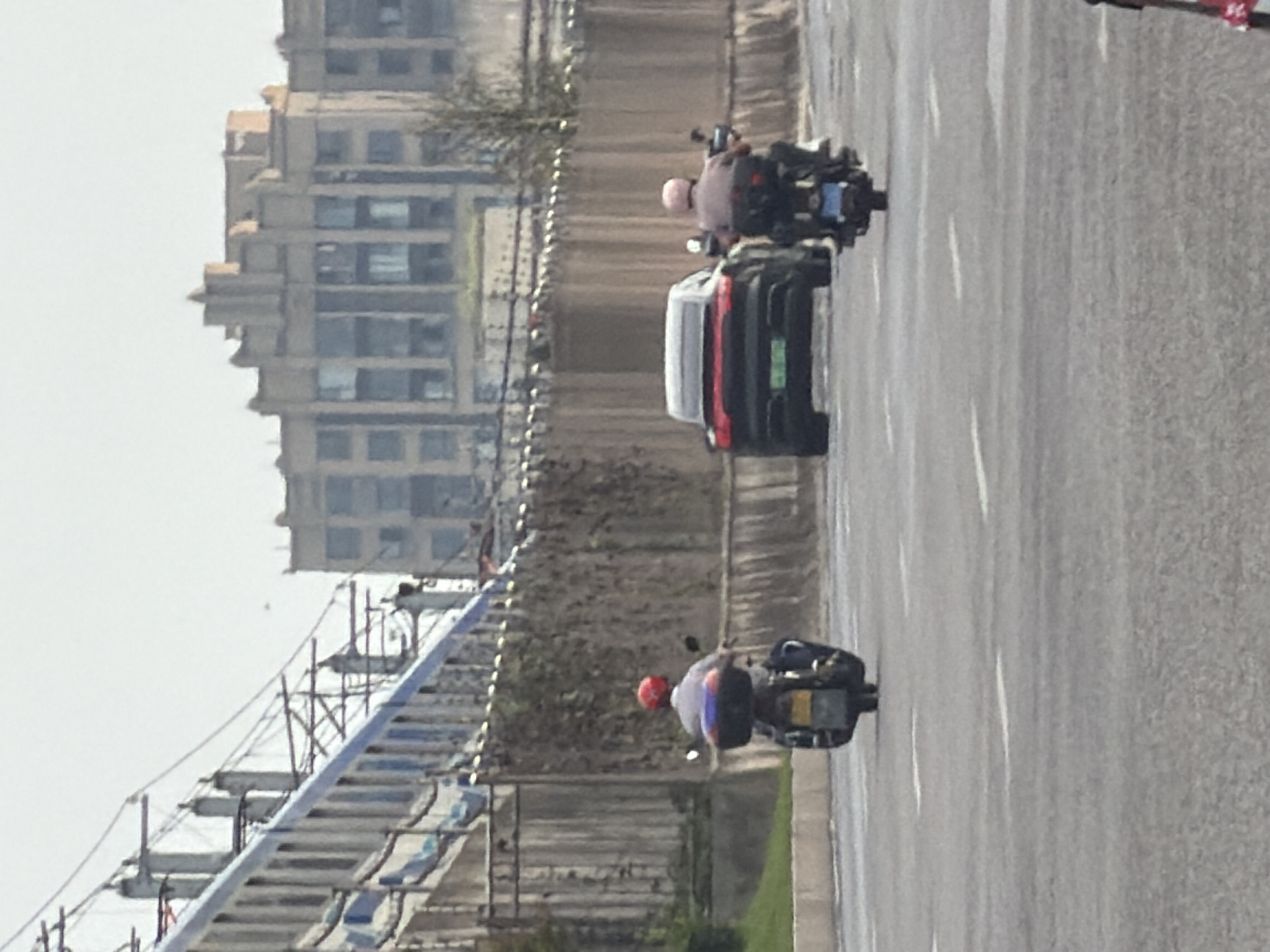 The image shows a view from a street in Jiangjin District, Chongqing, China, looking towards a road with several vehicles. In the foreground, two people are riding on motorcycles, one with a red helmet and the other with a pink helmet, both facing away from the camera. A dark-colored car, possibly a Porsche, is in the middle distance. In the background, a train travels along an elevated track, and beyond that, a multi-story building with distinctive architectural features is visible. A wooden fence runs along the side of the road, partially obscured by vegetation. The sky is overcast.