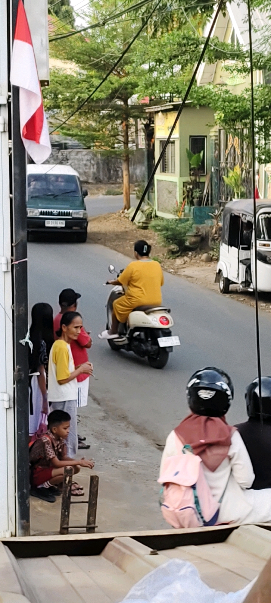 The image depicts a street scene in Makassar, Indonesia. The foreground shows the back of a boat-like structure, possibly a truck bed, with a piece of white plastic sheeting. To the left, a person wearing a brown and patterned school uniform and red shorts crouches near a wooden structure that resembles a ladder. Behind them, a group of four people stand on the sidewalk. The leftmost person is a young woman with long black hair, wearing a white and black patterned top and white pants. Next to her is a person wearing a red shirt and white shorts, with a black hat. A woman in a yellow shirt and grey pants stands with her head turned to the side, holding a cigarette. Further down the street, a person in a yellow outfit rides a beige scooter. In the background, a green van is driving away, and a white tricycle-like vehicle is parked. Several trees and buildings line the street, and power lines are visible overhead. An Indonesian flag is also visible on the left side of the image.