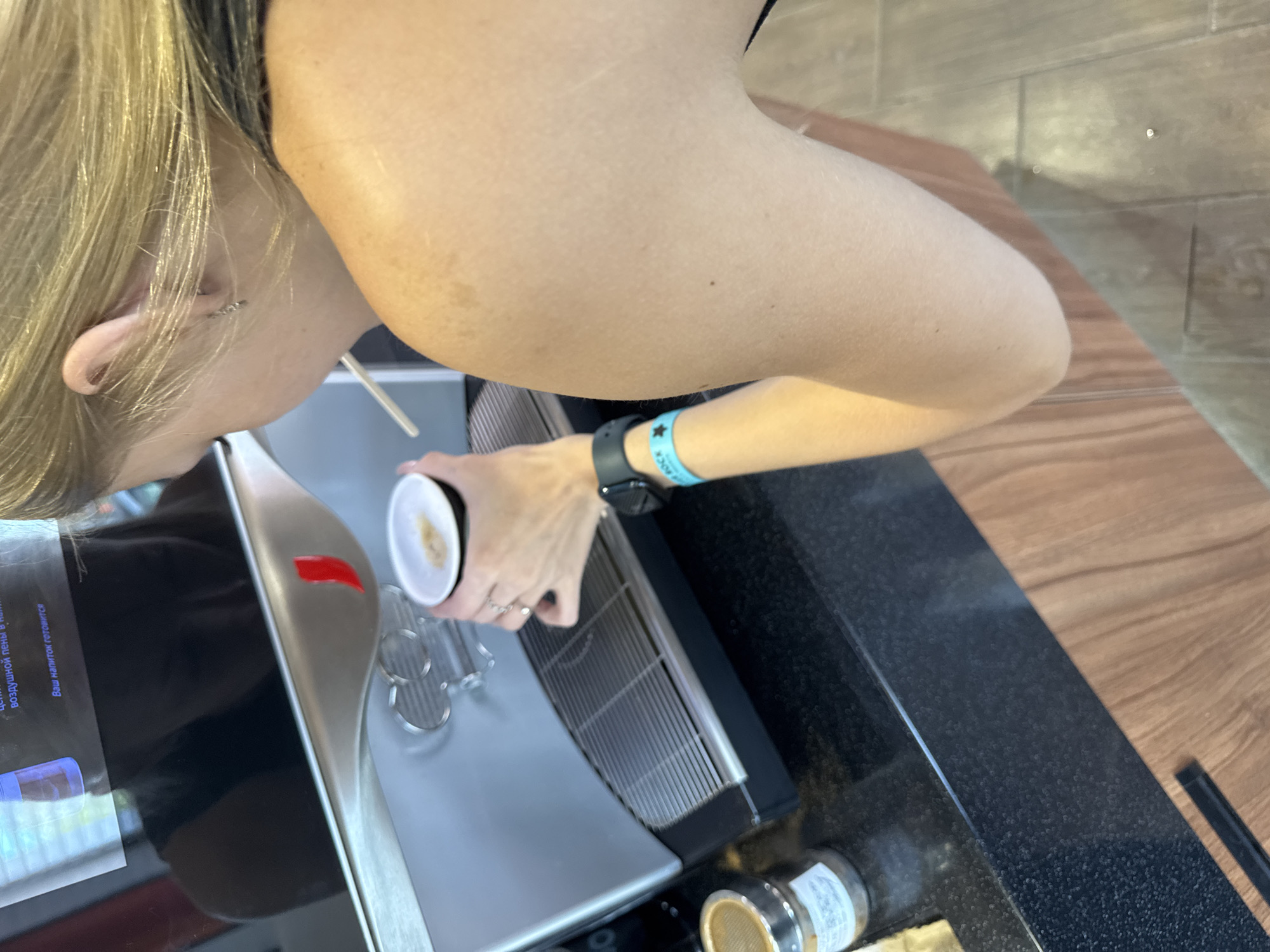 In Marchugi, Russia, a woman with blonde hair is standing at a coffee machine, preparing a beverage. She is holding a small cup with a white lid and a white straw. The coffee machine is silver and has a digital display that appears to be in Russian. The woman is wearing a black tank top and a dark watch on her left wrist. She also has a blue wristband on her left arm with the word "ROCK" printed on it. The background shows a dark countertop and wooden cabinets.