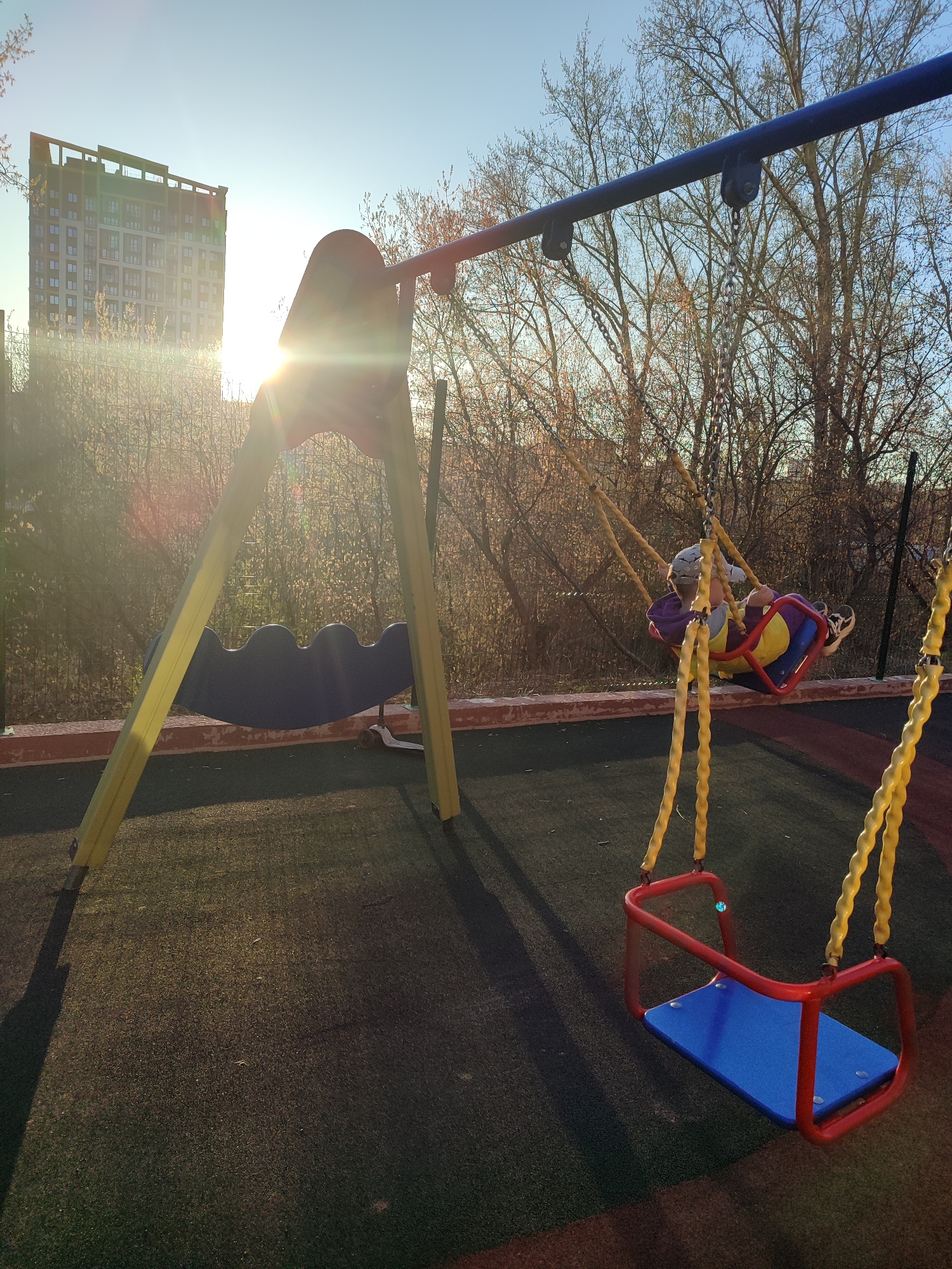 The image depicts a playground setting in Izhevsk, Russia. A child is swinging on a swing set. The playground features a swing set with a standard swing and a toddler swing. A building is present in the background. The environment is outdoors, with a rubber mat flooring. The image is taken during daylight hours, with the sun shining in the background.