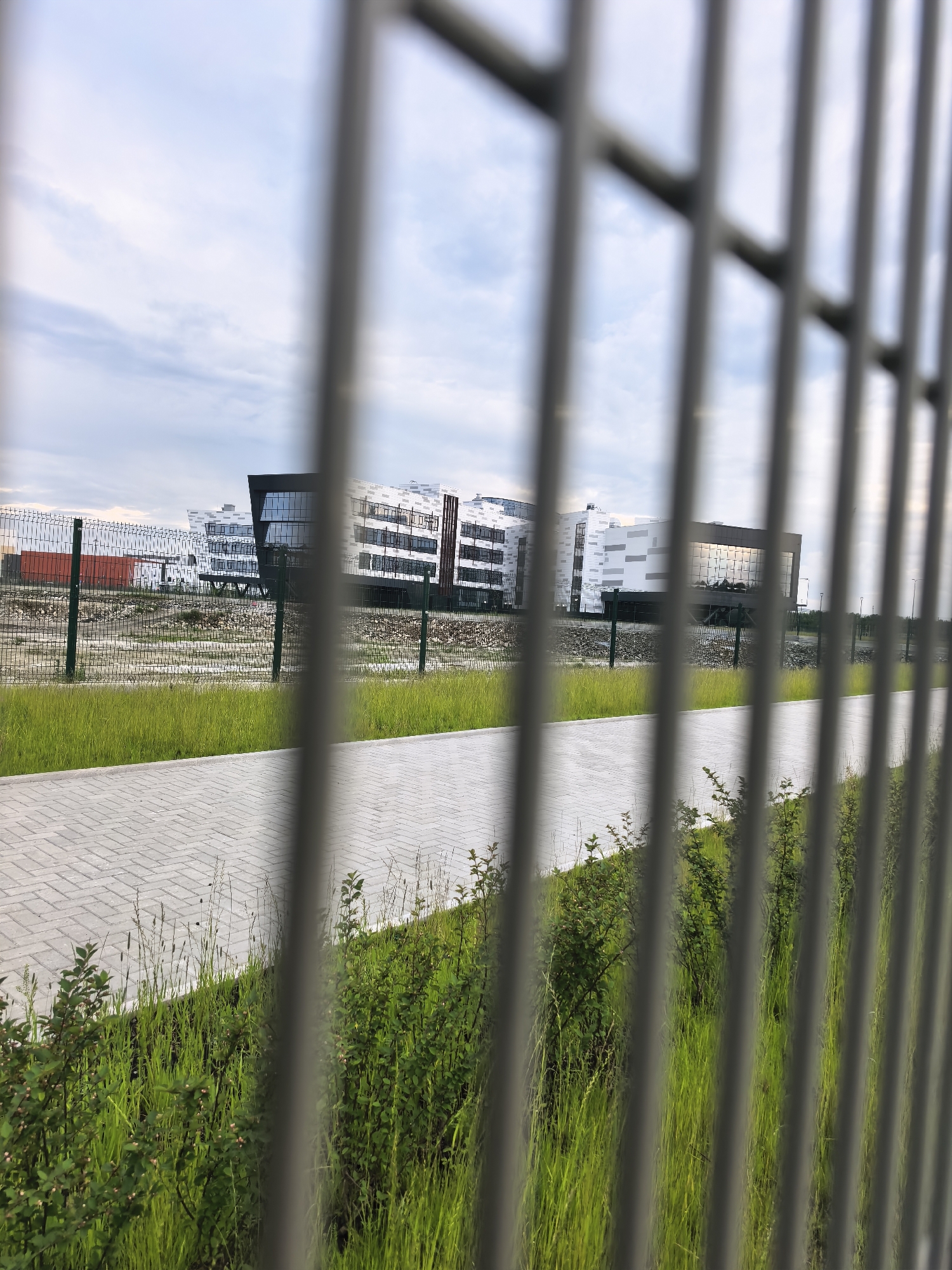 The image shows a modern, multi-story building complex with a reflective glass facade, viewed through a metal fence. The building is white and gray with geometric accents and appears to be in an industrial or business park setting. In the foreground, there is a paved walkway and lush green grass with some low-lying bushes. The sky is overcast with clouds. The scene is static with no people or animals visible.