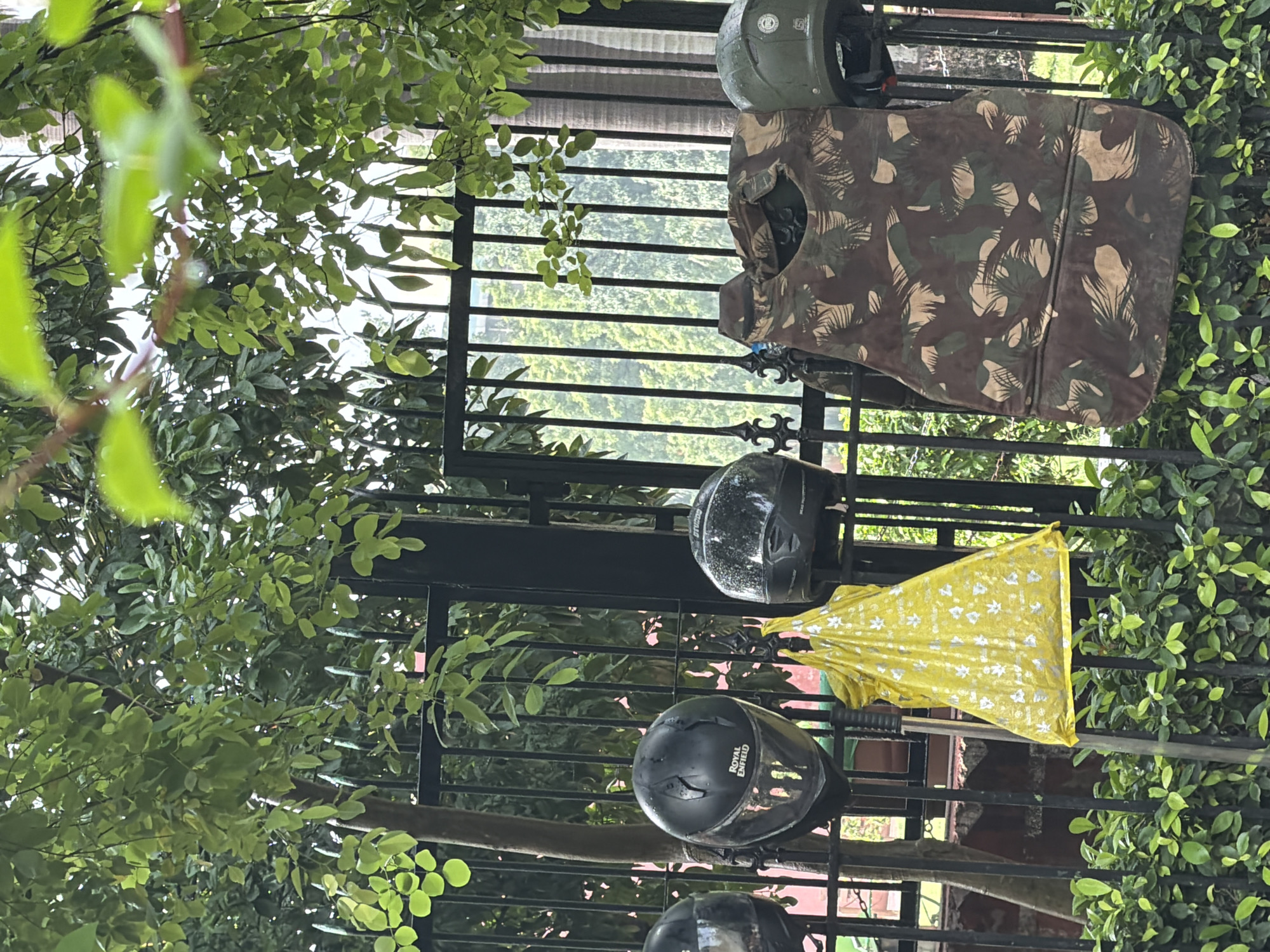 The image shows a black metal fence with a camouflage vest hanging on it, along with four helmets and a yellow bag. The camouflage vest appears to be a bulletproof vest, with a dense pattern of green, brown, and tan colors. Two of the helmets are black, and one is green. The yellow bag has a leaf pattern printed on it. Behind the fence, lush green trees and foliage are visible, suggesting an outdoor setting, possibly a park or a residential area. The presence of the vests and helmets could indicate a security checkpoint or a training exercise. The overall scene appears to be a roadside or a perimeter fence in New Delhi, India, given the context of potential security arrangements in a city of this size and type.