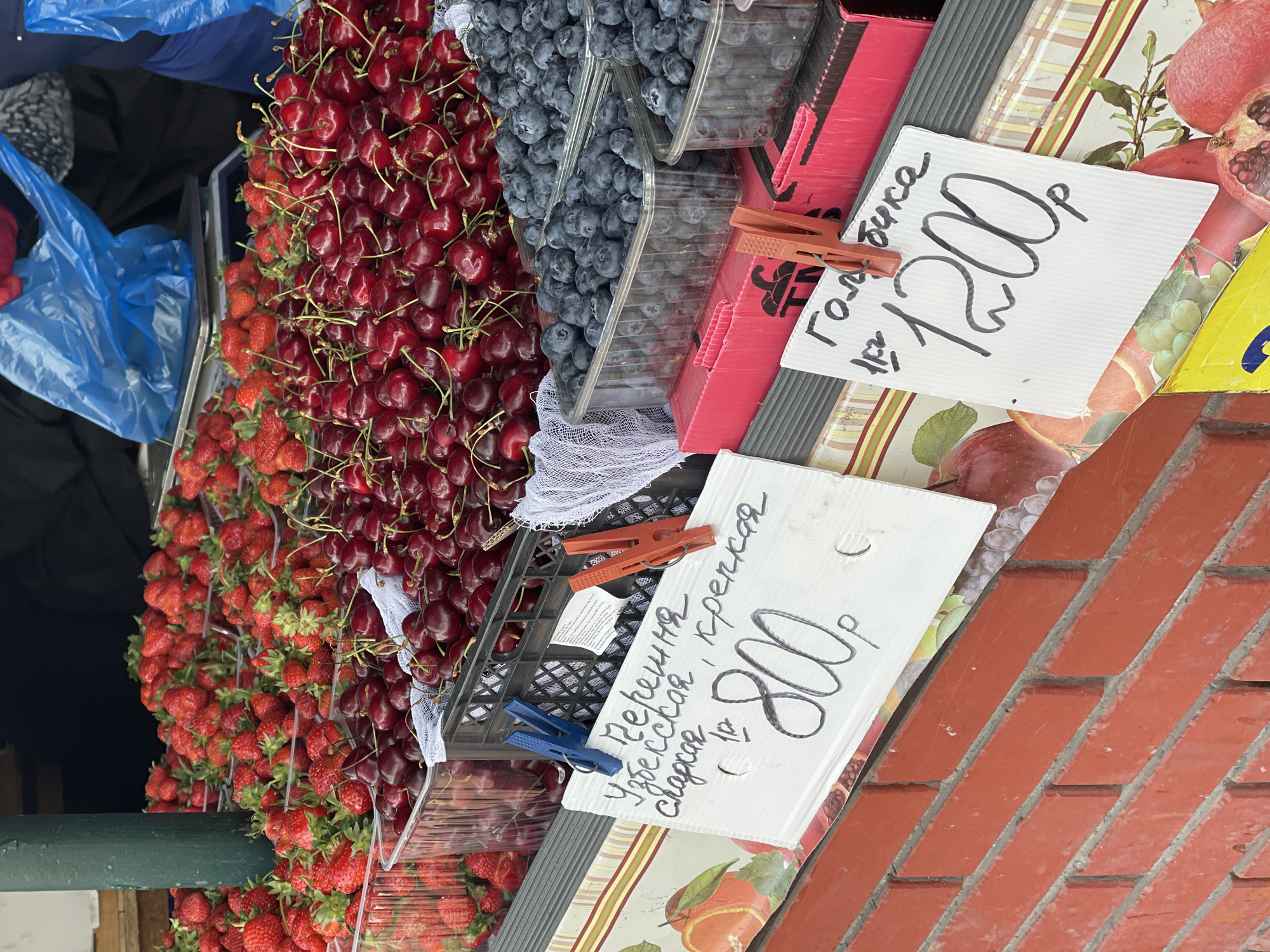 The image shows a fruit stand in the city of Unknown, Unknown. The stand offers strawberries, cherries, and blueberries displayed in clear plastic containers. Price signs are attached to the containers with clothespins; one reads "черешня Узбекская крепкая сладкая 800p", and the other reads "Голубика 1200p". The stand's surface is decorated with a fruit-themed pattern. A brick wall is visible to the side of the display.