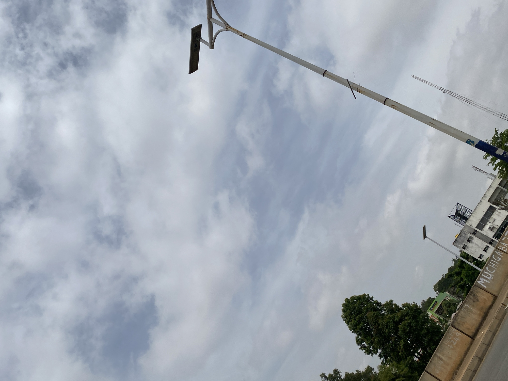 A tilted, low-angle shot captures a cloudy sky above a city street in Jos, Nigeria. In the foreground, a concrete barrier runs diagonally, displaying graffiti that includes the words "MUCHIGA BAZ." A grey paved road is visible to the left of the barrier. Beyond the barrier, a large green tree stands tall, with more trees and buildings in the background, including a white building with a blue lattice structure on its roof. A street light with a solar panel is prominently featured on the right side of the frame, leaning to the left. Another street light with a solar panel is visible further back, to the left of the prominent tree. The sky is filled with a mix of light and dark grey clouds, creating an overcast atmosphere.