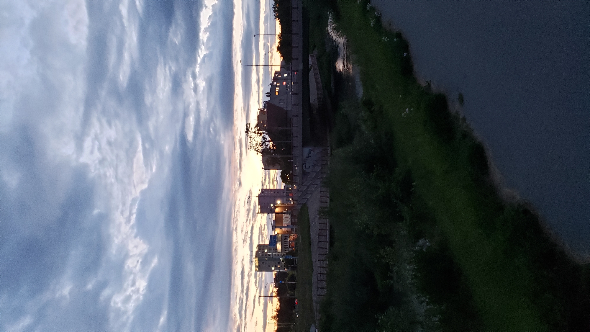 The image depicts an urban scene at dusk in Prešov, Slovakia, likely near the area of Sídlisko Sekčov. A body of water, possibly the Sekčov stream, flows through the foreground, bordered by lush green vegetation on the left bank. On the right bank, a paved pathway or road runs parallel to the water.
Dominating the midground is a concrete bridge spanning the water, with a pedestrian walkway and railings. Graffiti is visible on the underside of the bridge. Beyond the bridge, a collection of multi-story buildings forms the urban skyline. These structures appear to be residential or office buildings, with some windows illuminated by interior lights, suggesting it is late evening. Streetlights are also visible, casting a dim glow. The sky above is filled with dramatic, cloudy formations, with hints of the setting sun creating a soft, warm light on the horizon. The overall atmosphere is calm and serene, characteristic of twilight in a city. No people are visible in the scene.