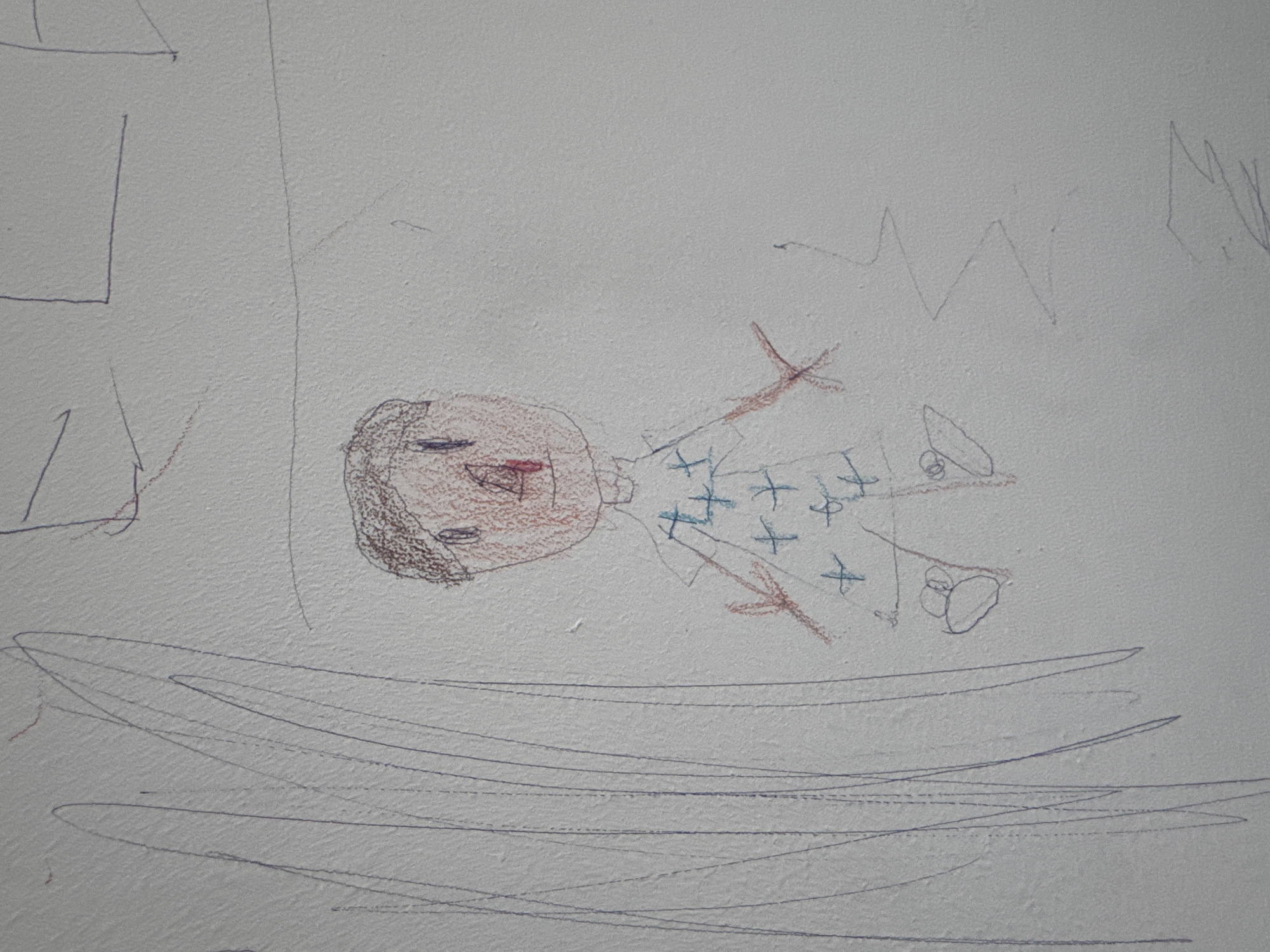 The image displays a child's drawing, primarily featuring a stylized human figure, executed with crayons or colored pencils on a light-colored, textured surface, possibly a wall or thick paper. The main figure is drawn in the center, oriented sideways, with its head to the left and feet to the right.
The person depicted has a round, reddish face with simple blue lines for eyes and a small red mark for a mouth. Dark brown scribbles outline the head, possibly indicating hair. The body is rectangular, colored light blue, and adorned with multiple blue 'x' marks, suggesting a patterned outfit. Thin, reddish-brown lines form the limbs, and two small, dark-lined oval shapes are near the bottom, potentially representing feet or shoes.
Around the central figure, various other drawings and scribbles are present. To the left, a partial dark blue rectangular outline and other faint lines are visible. Below the figure, several long, wavy, dark blue horizontal lines span the width of the lower part of the image, possibly depicting water, ground, or a bed. To the upper right, prominent zigzag lines in dark blue add an abstract element, perhaps representing mountains or an irregular pattern. There are also scattered faint lines and marks across the surface.
No discernible text is visible in the drawing. The overall impression is a vibrant and imaginative piece of art created with a child's uninhibited style.