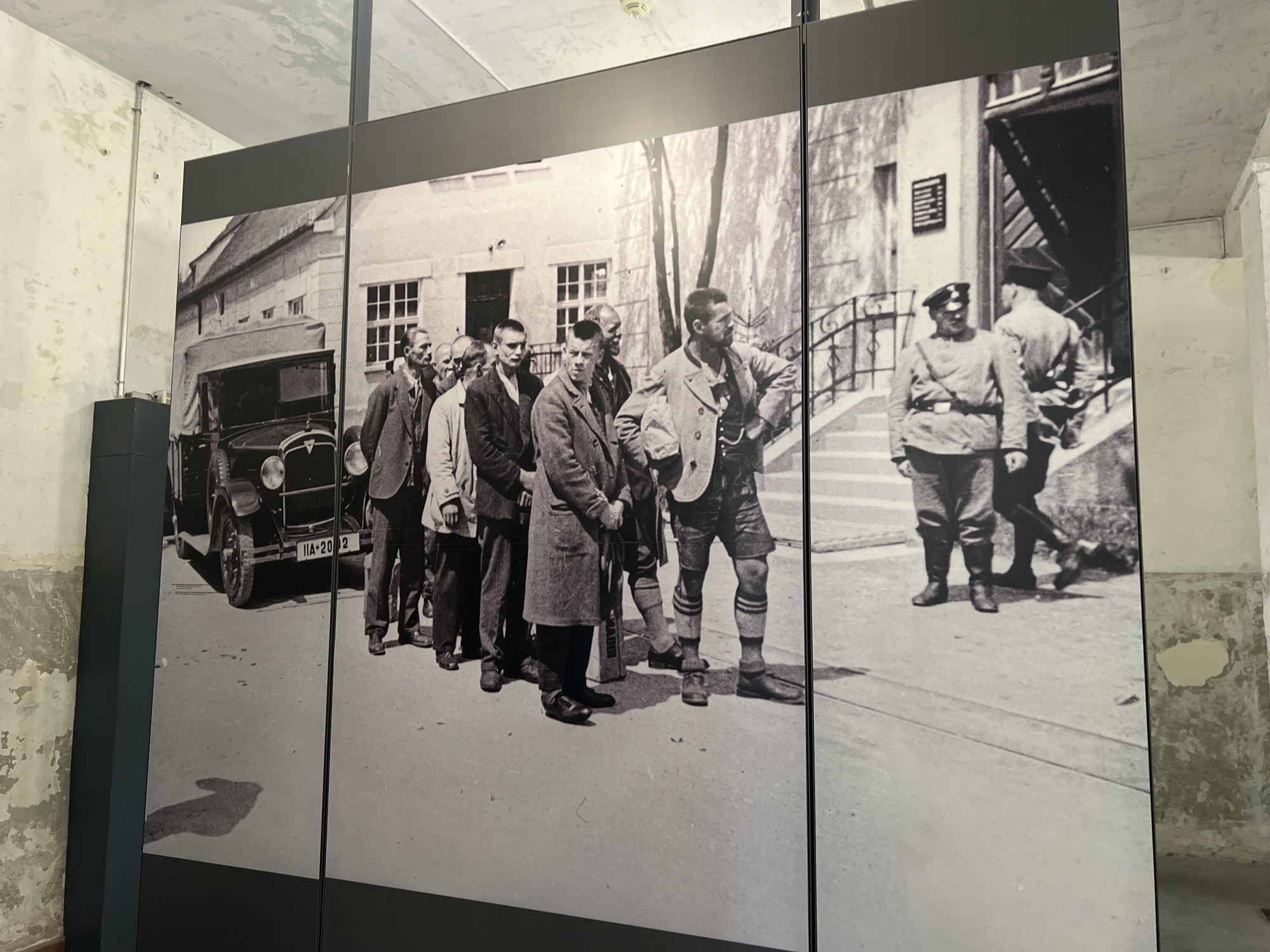 The image depicts a large black and white photograph displayed in a museum setting, likely a former prisoner barracks or an associated building within the Dachau Concentration Camp Memorial Site in Dachau, Germany. The photograph shows a group of men, some in civilian clothing and others in what appear to be uniforms, standing in a line on a paved area outside a building with an entrance and steps. A vintage automobile is parked to the left of the frame. The men are facing in various directions, with some looking towards the building and others looking away. The overall scene is grim and appears to document an event related to the history of the concentration camp. The surrounding environment within the museum shows rough, unpainted concrete walls and ceiling.