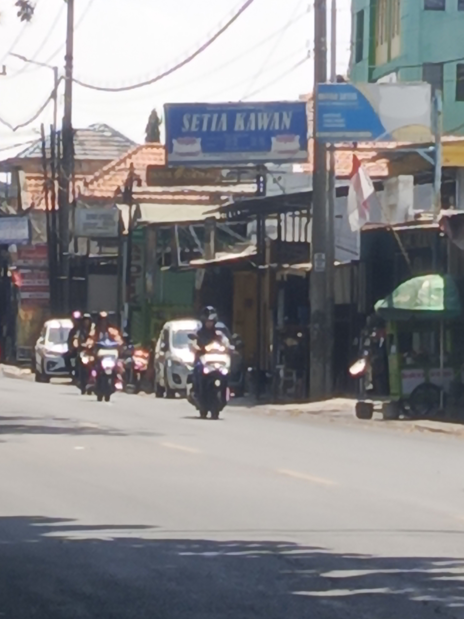 The image depicts a street scene in Kedungrejo, Indonesia, on a sunny day. In the foreground, a paved road is visible, with shadows cast by unseen objects or trees on the left side. Several motorcycles are traveling on the road, with their headlights illuminated. Two white cars are parked or moving in the background. Buildings line the right side of the street, including a multi-story structure with green facades and a smaller building with a tiled roof. A blue billboard with white text, partially obscured, is prominently displayed above some of the buildings. On the right side of the street, a green stall with an umbrella is set up, possibly for selling goods. The overall impression is of a bustling urban environment.