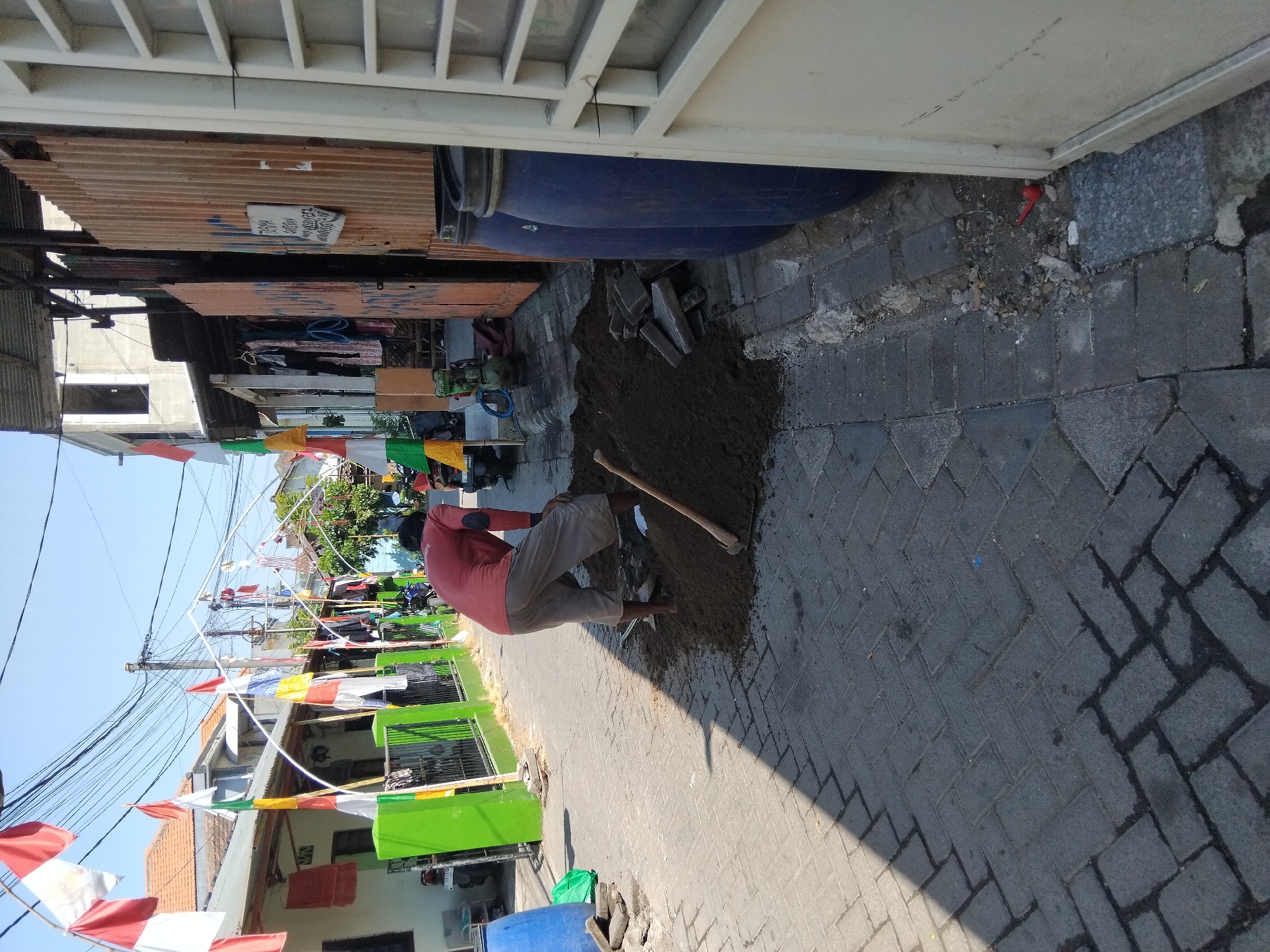 In Kepuh Kiriman, Indonesia, a man wearing a red long-sleeved shirt and gray shorts is shoveling dirt from a pile on a paved walkway. The walkway is made of dark gray interlocking bricks. To the left of the man, there is a bright green fence and several Indonesian flags hanging above the walkway. To the right, there are two large blue barrels and a light-colored door. The scene appears to be a street or alleyway.