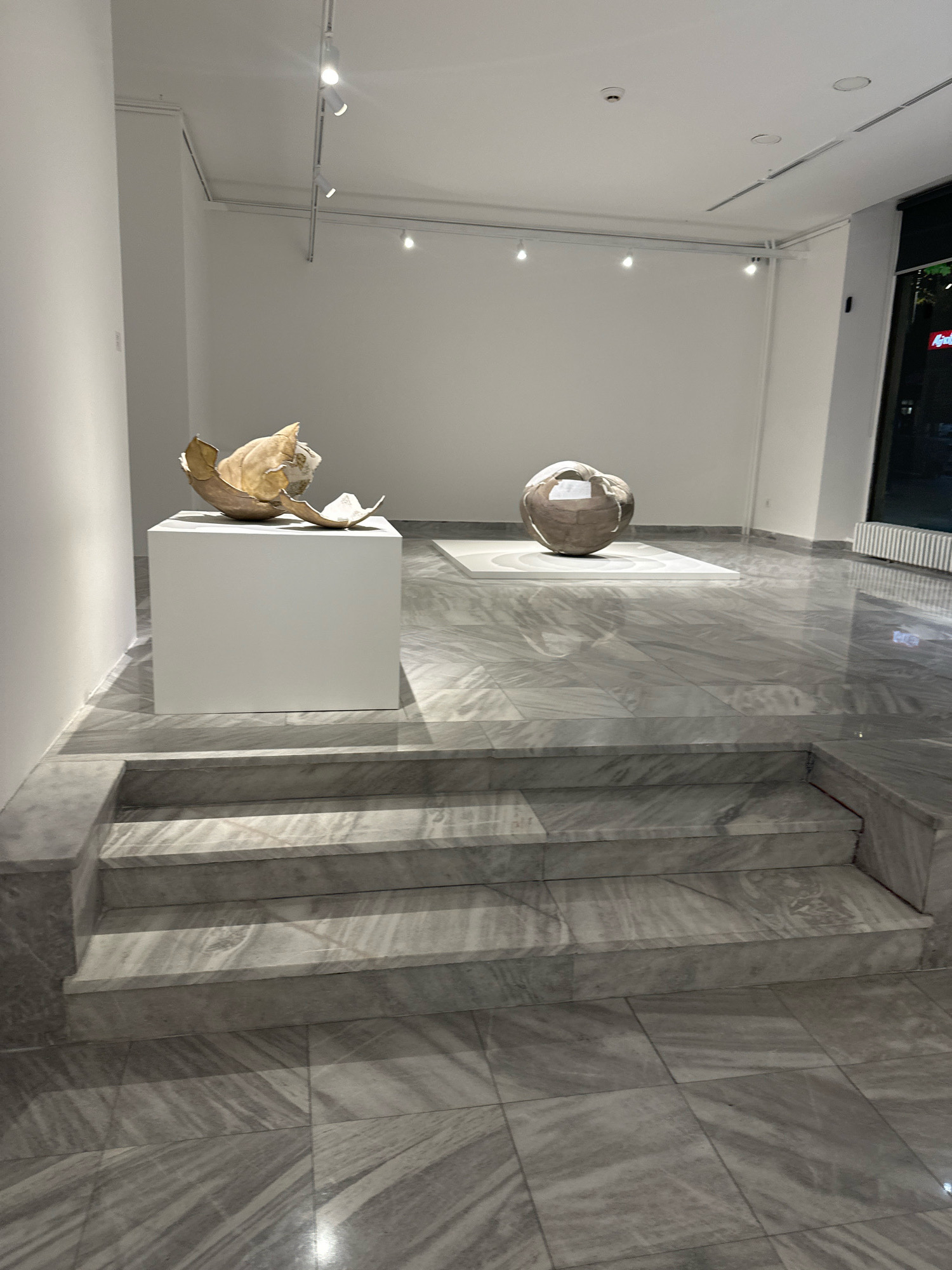This image depicts the interior of a modern art gallery in Belgrade, Serbia. The space features white walls and a polished grey marble floor with intricate veining. A set of marble stairs with subtle lighting leads up from the foreground. On the left, a white cubic pedestal displays a fractured, shell-like sculpture with a golden-brown interior. Further into the gallery, a second, larger spherical sculpture with a woven texture sits on a white rectangular platform. Track lighting is visible on the ceiling, illuminating the artworks. To the right, a large window reveals a darkened exterior with glimpses of neon lights. The overall ambiance is minimalist and sophisticated, typical of contemporary art exhibition spaces.