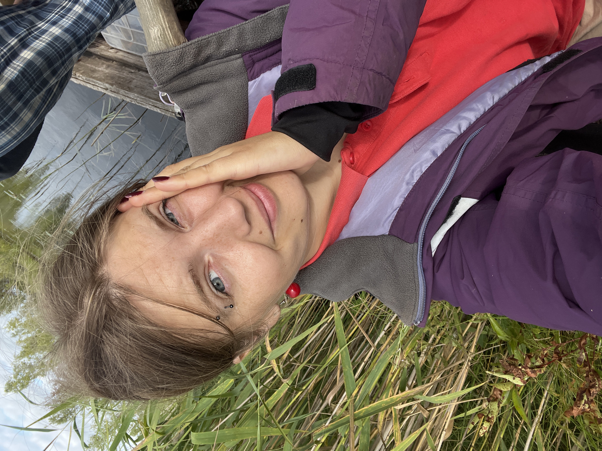 The image shows a close-up, selfie-style shot of a young Caucasian woman with blue eyes, fair skin, and freckles, resting her head on her right hand. She has brown hair, styled casually, and piercings above her right eyebrow. She is wearing a red polo shirt underneath a purple jacket with grey accents. The background consists of tall reeds and a body of water, suggesting an outdoor, natural setting. Part of another person, wearing a blue and grey plaid shirt, is visible on the right side of the frame, along with a wooden structure. The location is identified as Navasyolkaŭski sel'ski Save, Belarus.