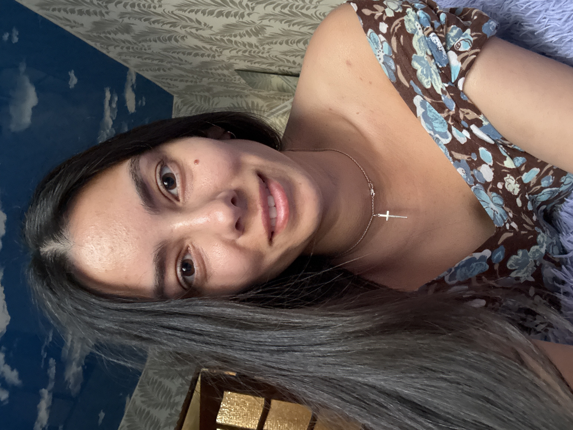 A woman with long dark hair, brown eyes, and a clear complexion is positioned in the foreground, partially turned towards the viewer. She is wearing a brown and blue floral dress and a silver necklace with a cross pendant. Her expression is a soft smile, and her gaze is directed towards the camera.
Behind her are two different wallpaper designs. The upper part of the background features a light gray wallpaper with an intricate, repeated pattern. The lower part shows a blue wallpaper printed with a cloudy sky scene.
No information is available to confirm the location in the city of Chaikovsky, Russia.