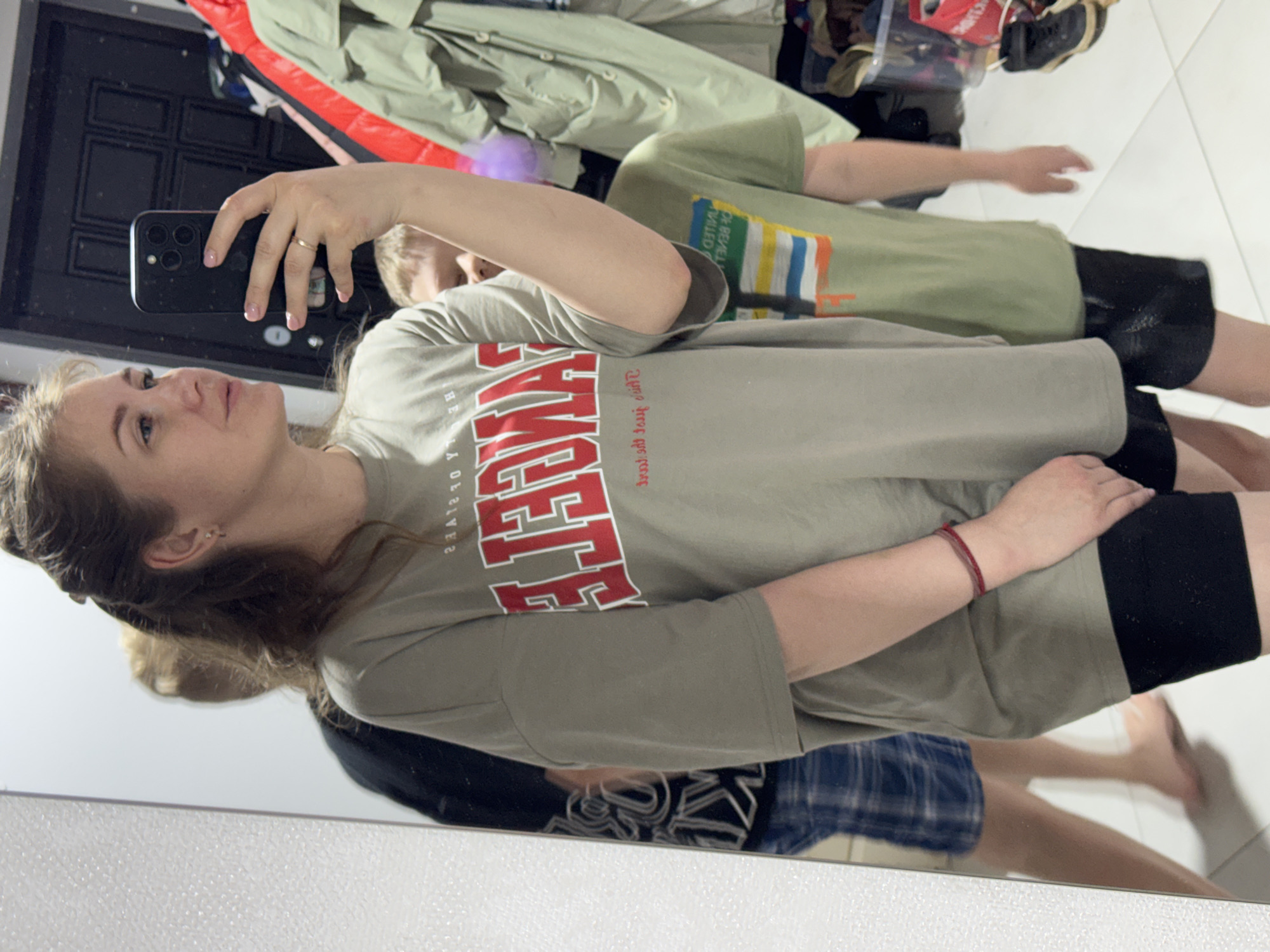 In a mirror's reflection, a woman with light skin and brown hair, parted to her left, is taking a selfie with a black smartphone held in her left hand. She wears a gray t-shirt with red lettering and black shorts. A gold ring is visible on her finger. Behind her, another person, possibly a child, is seen wearing a green t-shirt and black shorts. Additional people are visible in the reflection, one wearing blue plaid shorts and another wearing a black shirt. A variety of garments hang on a rack in the background. The scene appears to be indoors, possibly in a dressing room or bedroom. It is impossible to determine the precise location in Gornyi (Dvurechenskoe S/P), Russia, from the provided image.