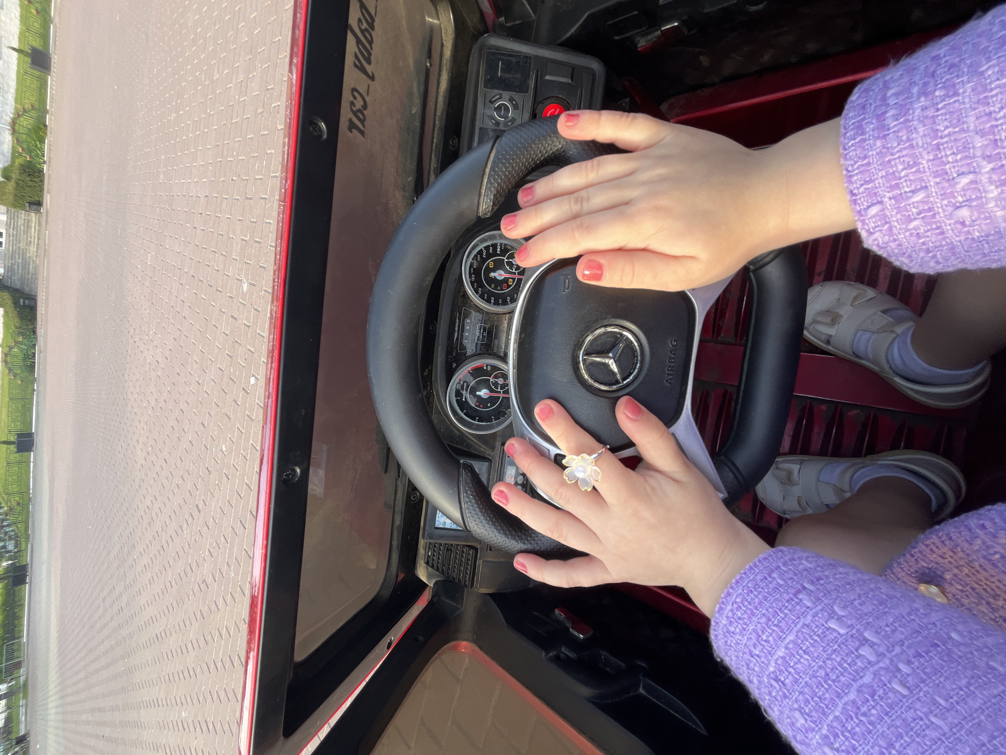 A top-down, slightly angled view captures a person's hands and lower body inside what appears to be a child's electric ride-on car. The person's hands, with red nail polish, grip a black steering wheel that prominently displays a silver Mercedes-Benz logo and the word "AIRBAG." A flower-shaped ring adorns the ring finger of the right hand.
The individual is dressed in a textured, light purple long-sleeved top or jacket, white socks, and light-colored sandals. Beyond the steering wheel, a toy dashboard features mock gauges, including a speedometer with markings like 20, 40, 60, 80, and a fuel gauge with "F/E." Gear indicators "P R N D" and various control buttons, including a red power button, are visible. The car's interior elements are red and black. To the left of the steering wheel, text resembling "pspa_csr" and "163_vdga" is inscribed on a dark panel.
The vehicle is positioned outdoors on a wide expanse of light-colored, meticulously laid interlocking brick pavers that stretch into the distance. In the far background to the left, glimpses of greenery and what appears to be a low fence or barrier can be seen. The bright, even illumination suggests the photograph was taken during daytime. The image depicts the activity of a person, likely a child, engaging in play by "driving" the toy car.