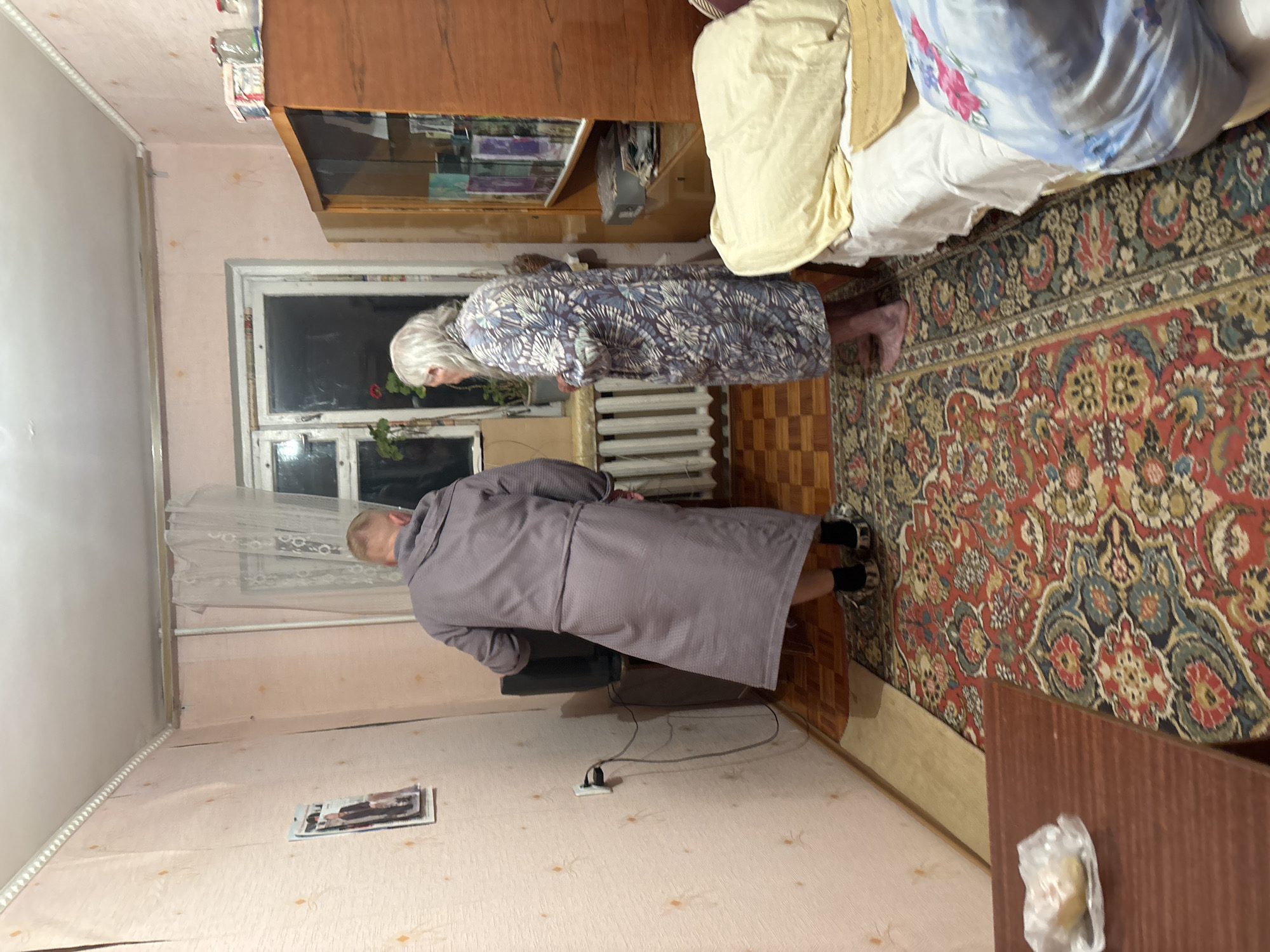 Two individuals are present in a room. One, an elderly woman with grey hair, is wearing a patterned robe. The other, a shorter individual with blonde hair, is wearing a purple robe. The room is furnished with a bed, a wooden cabinet, a radiator, and a rug. The wall is pink, and a window is visible. A magazine rests on the floor. The scene appears to be indoors. No location information is specified.