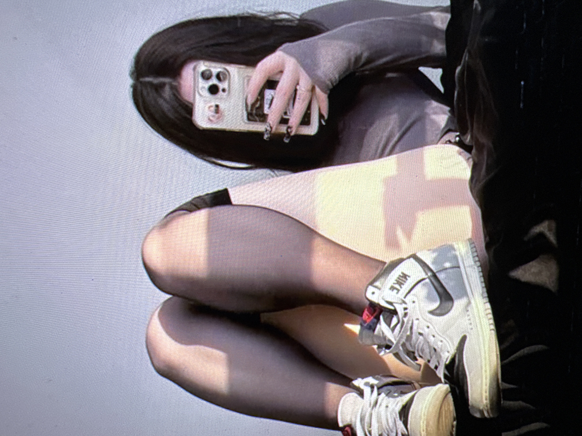 The image shows a person's legs and torso, with a focus on their footwear. The person is wearing black pantyhose and white Nike high-top sneakers. The sneakers have a black Nike swoosh logo and the word "NIKE" on the heel. The person is also holding a smartphone in their right hand, which is partially visible. The phone has a silver case with multiple camera lenses. The background is a light gray surface with faint horizontal lines. Sunlight is casting shadows on the person's legs, suggesting it is daytime. The overall impression is a casual, self-taken photograph.