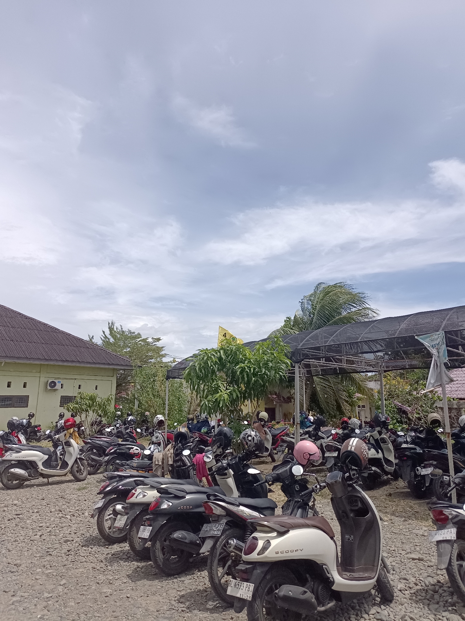This is a medium-angle, eye-level, full shot of a large outdoor parking lot filled with many motorcycles. The ground is covered with gravel. In the background, there is a light green building with a dark gray roof, and behind that, dense green trees and vegetation. A dark-colored canopy with a netted covering is visible in the middle ground. The sky above is partly cloudy with a mix of white and gray clouds. Several motorcycles are parked close together, with some helmets on their seats or handlebars. One white scooter in the foreground has the word "SCOOPY" visible on its side. Another motorcycle in the foreground has a license plate that reads "BL 6533 PBJ". The overall scene depicts a busy parking area, likely in a tropical or subtropical climate given the vegetation and cloud cover.