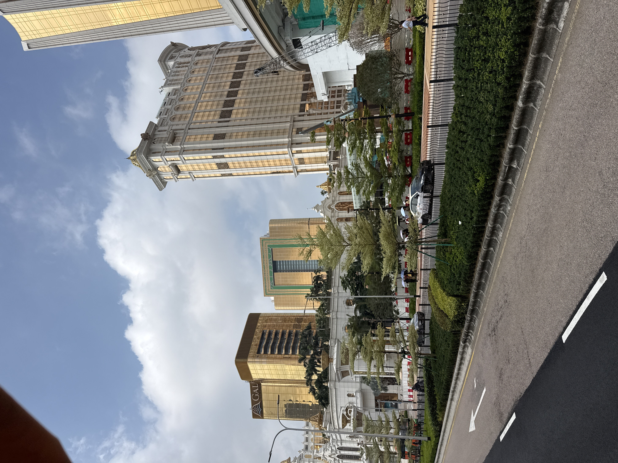 The image depicts a street scene in Unknown, Macao, featuring a road with an arrow marking indicating traffic direction. Adjacent to the road is a line of bushes and a fence. Behind the fence, there are several buildings, including high-rise structures with golden facades. The sky is visible above the buildings, with a mix of blue and white clouds. The architectural style of the buildings suggests a modern, possibly commercial or residential area.