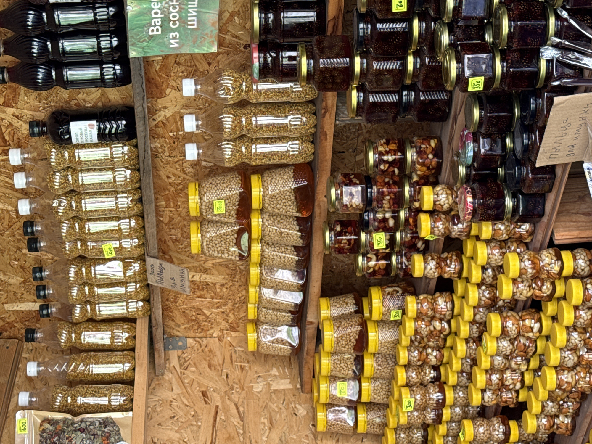 This image depicts a stall in Chemal, Russia, likely a market or roadside vendor, selling various preserved goods. The stall is constructed with wooden planks, suggesting an outdoor or rustic setting.
The primary subjects are rows upon rows of glass jars and plastic bottles filled with food items. The jars vary in size and shape, many with yellow lids, and contain what appears to be nuts, dried fruits, or seeds immersed in a liquid, possibly honey or syrup. Some jars are stacked in multiple tiers, filling the shelves. There are also several clear plastic bottles filled with small, golden granules, possibly bee pollen or seeds.
On a higher shelf, dark glass bottles are visible, some with black caps and others with dark liquid. A green sign with white text, likely a price or product label, is positioned to the right of these bottles. A small, brown cardboard sign with handwritten text in Cyrillic is placed on a lower shelf.
The overall impression is one of abundance and variety, with a focus on natural, possibly homemade, products. The lighting suggests it is daytime, with diffused light illuminating the scene. No people are visible in the image, but the arrangement of goods implies a vendor setting.