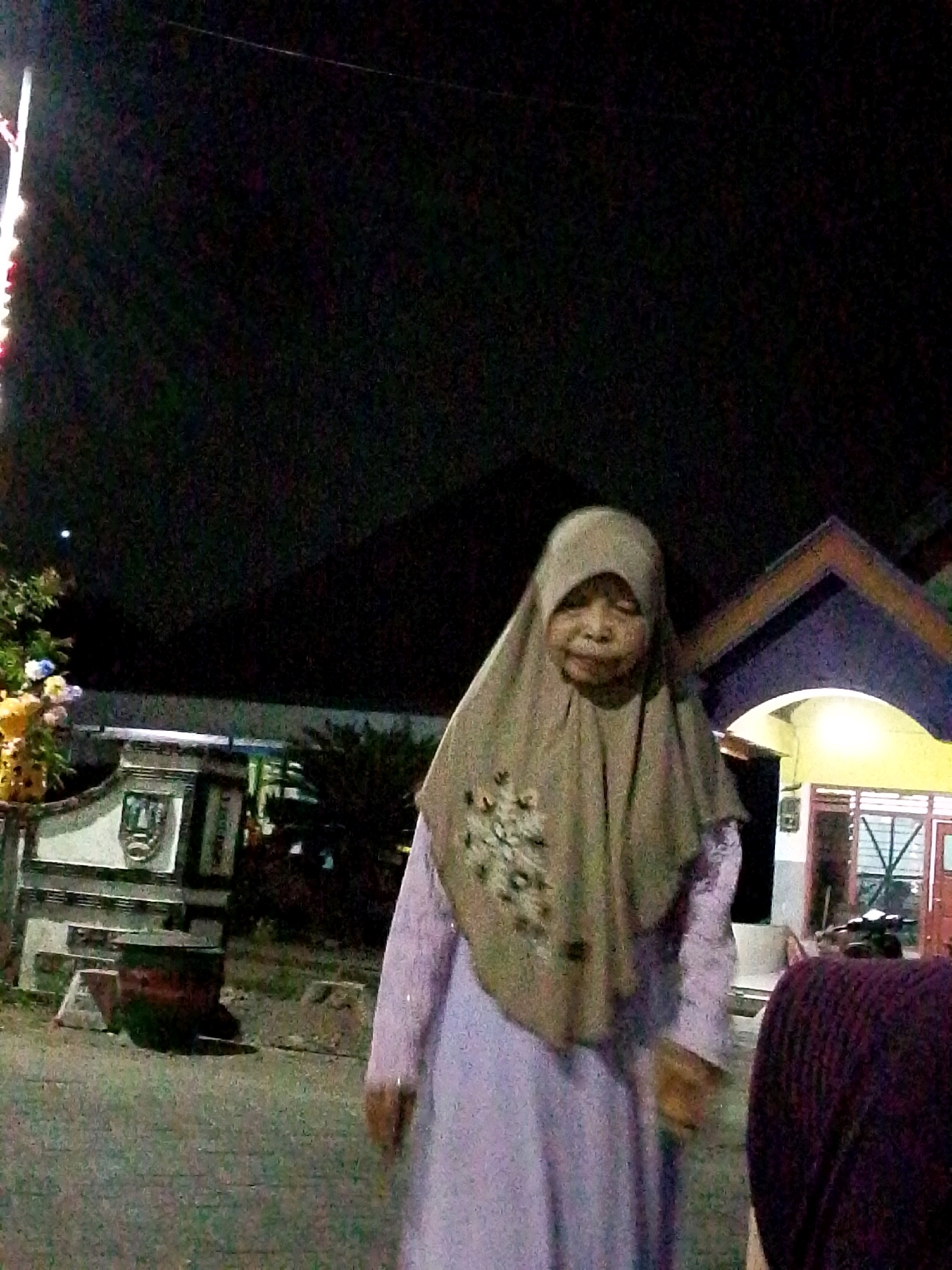 An elderly woman wearing a light brown hijab and a long light purple dress stands in front of a building with a yellow facade and a dark purple roof. She is facing slightly away from the camera. Behind her, to the left, is a white structure with decorative elements and what appears to be a crest or emblem. To the far left, a light pole with red and white lights is visible. The ground is paved with cobblestones. The scene appears to be outdoors at night, as indicated by the dark sky and the lighting from the building. The location is identified as Bulurejo, Indonesia.