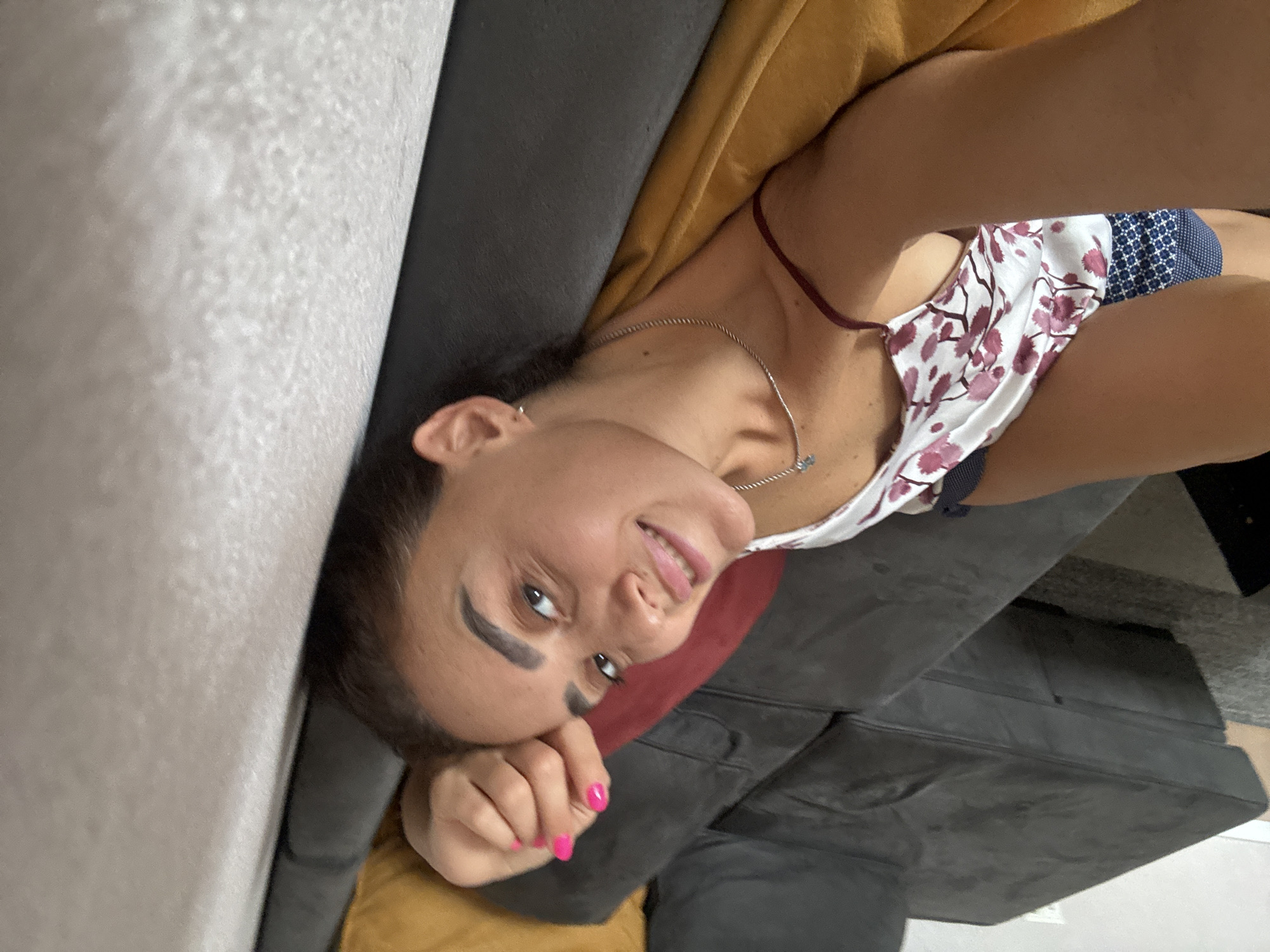 A young woman is lying on a sectional sofa in a room. The sofa is dark gray and has a mustard yellow pillow visible. The woman has dark hair, brown eyes, and is wearing a white top with a floral pattern. She has bright pink painted fingernails. The woman is looking at the camera and smiling. The scene appears to be indoors, possibly in a living room. There are no specific landmarks visible to suggest the location is in Tbilisi, Georgia.