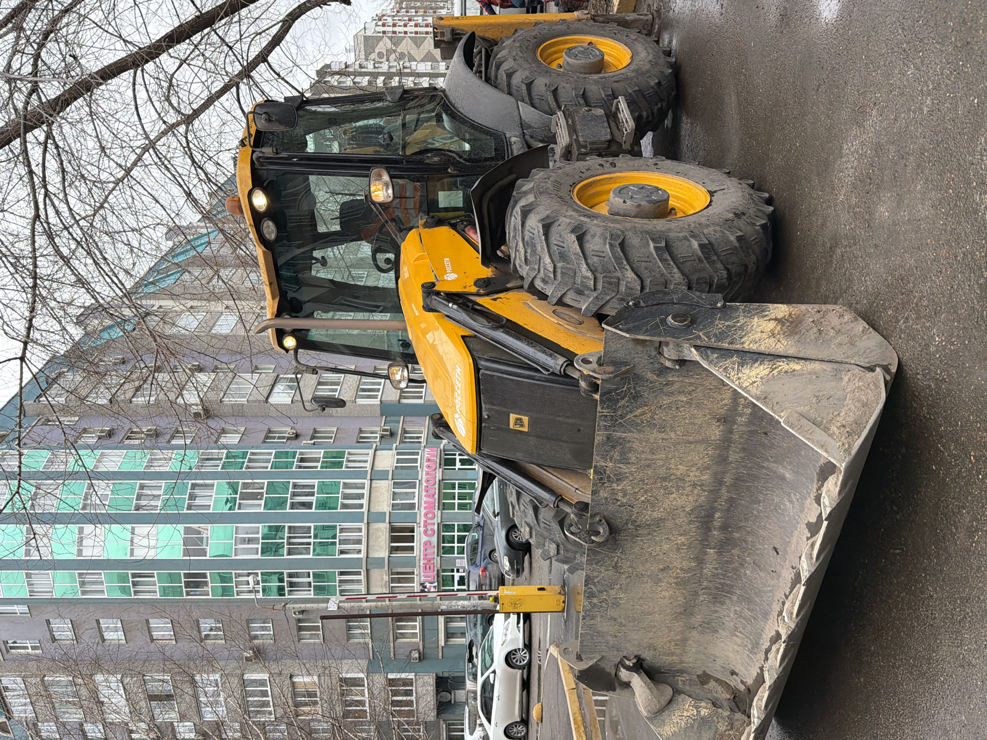 The image shows a yellow backhoe loader parked on a street in Krasnoyarsk, Russia. The backhoe is facing to the right. Behind it is a multi-story building with a sign in Russian, "ЦЕНТР СТОМАТОЛОГИИ" (Dentistry Center). A parking gate and several cars are also visible behind the backhoe. Bare tree branches are visible in the foreground. The ground appears to be wet.