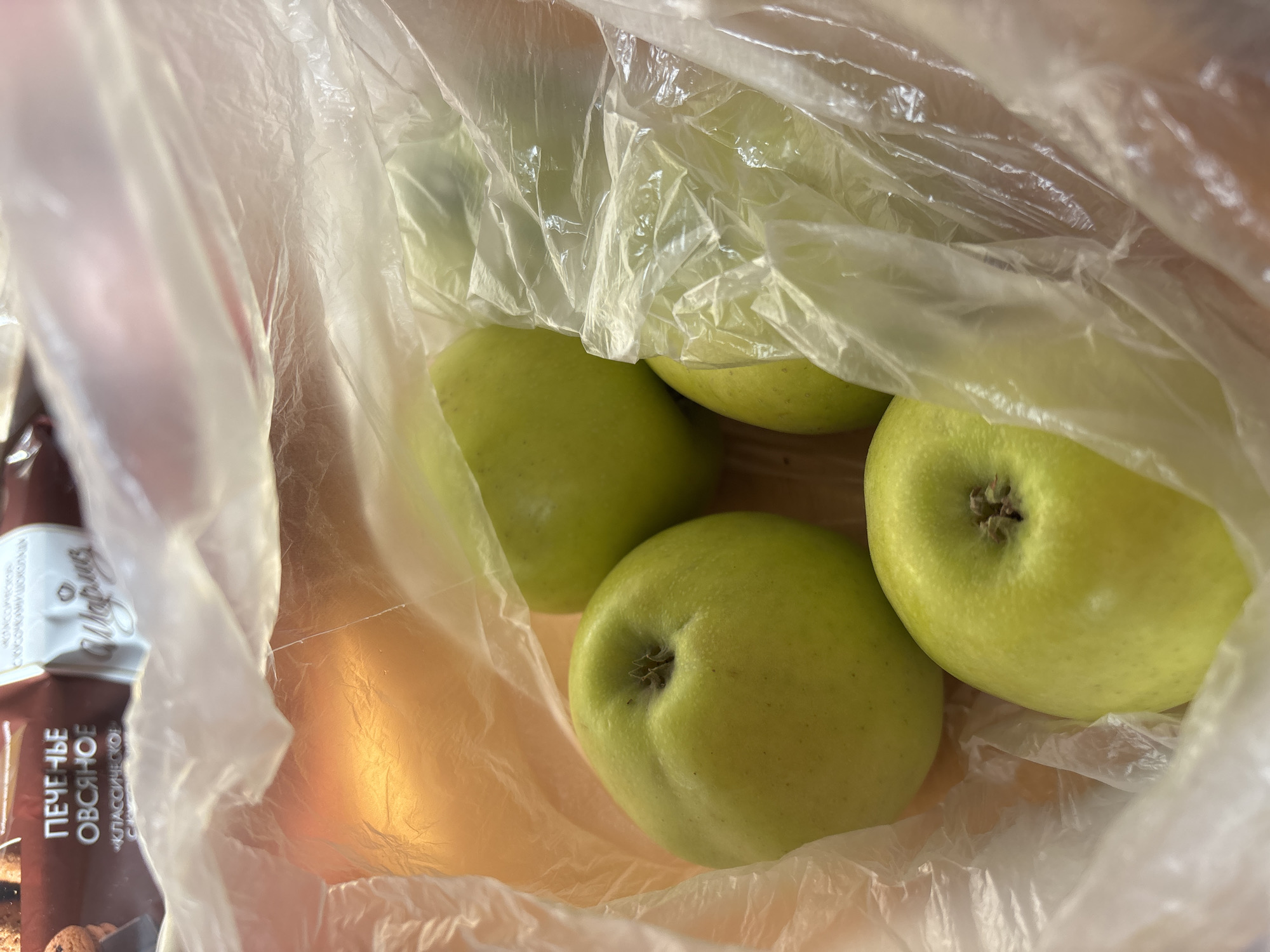A slightly high-angle view depicts a translucent plastic bag containing several green apples. The bag is partially obscuring a brown package of what appears to be cookies or crackers, labeled in Russian with "ПЕЧЕНЬЕ ОВСЯНОЕ" (Oatmeal Cookies) and "КЛАССИЧЕСКОЕ" (Classic). The background appears to be a plain, light-colored surface. Location is unknown beyond what is contained in the image. The city of Vsevolozhsk, Russia is not apparent in the image.