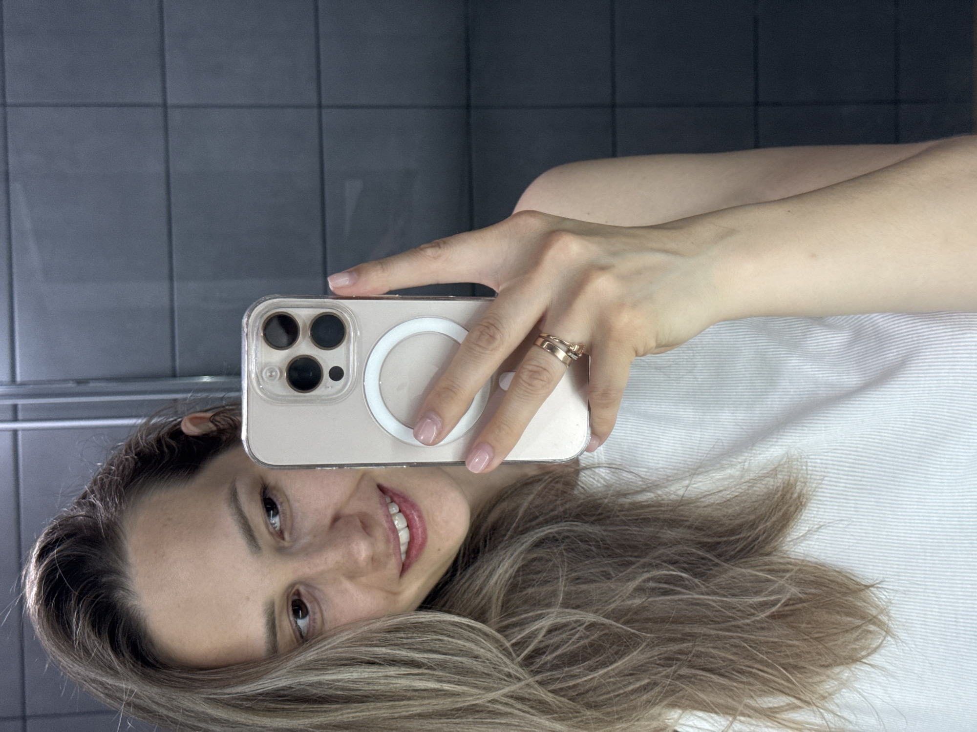 A selfie shot captures a woman with light brown and blonde hair, smiling slightly. She holds a smartphone, likely an iPhone, in a clear case with a visible MagSafe charging ring on the back. Two gold rings are visible on her fingers. The background suggests a bathroom setting with light grey tiles. No evidence indicates a specific location or city.