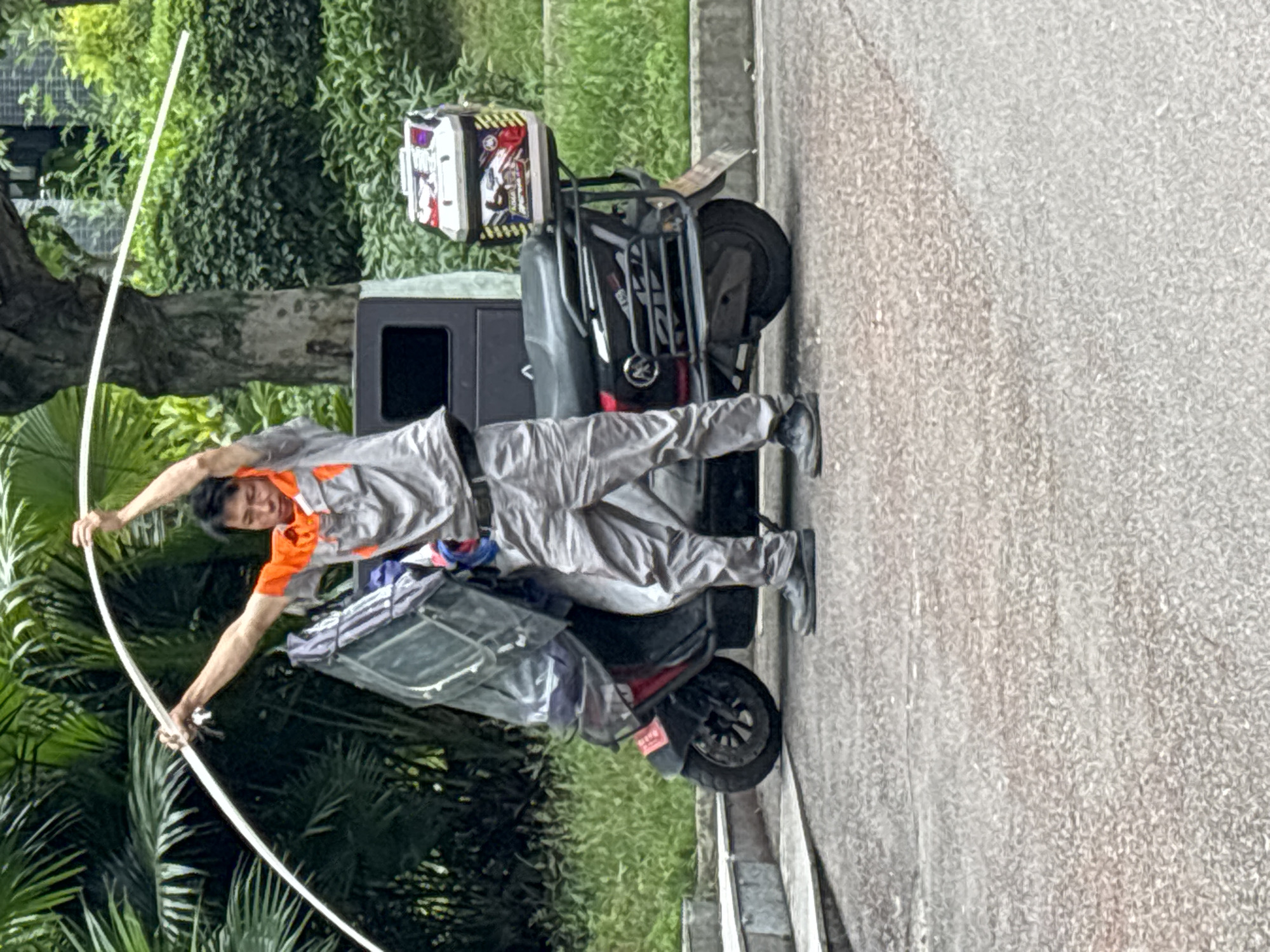 A man in a grey and orange work uniform stands next to a scooter on a paved surface, holding a long, curved white object, possibly a pipe or conduit, in both hands, raised above his head. The scooter has a black frame and a rear-mounted box. The background consists of trees and vegetation, suggesting a roadside setting. The location is Jiangjin, China.
