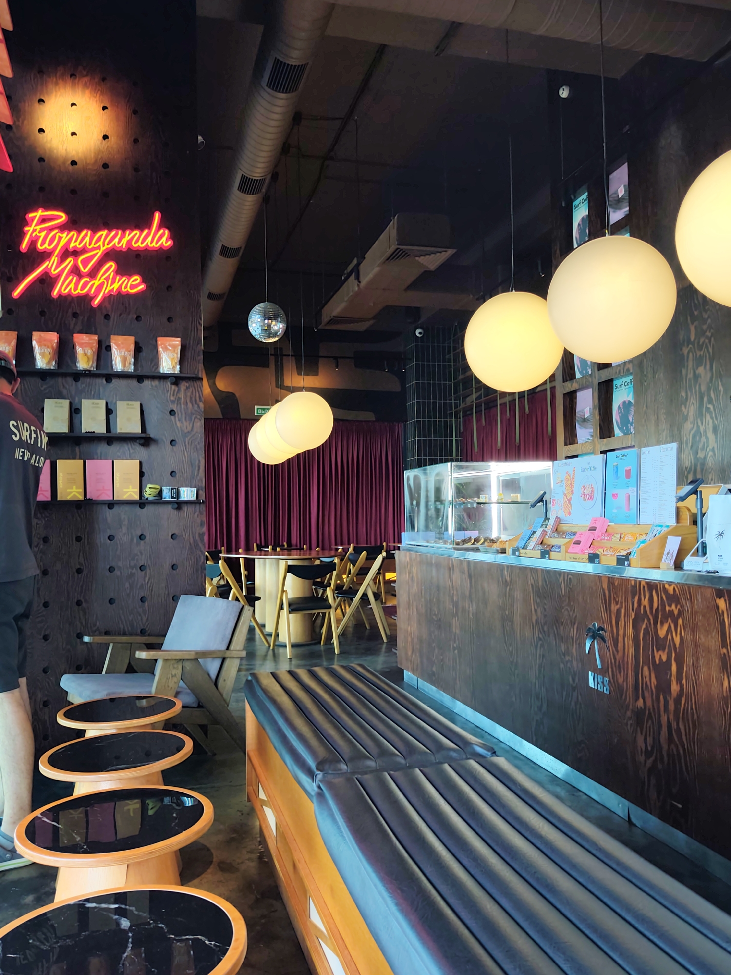 The photograph captures the interior of a modern cafe, potentially a branch of "Propaganda Machine" given the neon sign. The cafe is decorated with dark wood paneling and deep red curtains, creating a cozy yet stylish atmosphere. In the foreground, a long, black leather bench is partially visible, with a row of oval-shaped side tables with black marble tops next to it. The cafe features industrial-style ventilation ducts and a disco ball hanging from the ceiling. Several large, spherical lamps illuminate the space with a warm glow. On the left side of the image, shelves display packaged products, and a man wearing a dark t-shirt with "SURFING NEW ALO" is standing. The counter area displays a glass case with pastries and various menus. The overall ambiance suggests a contemporary urban eatery. While the image itself does not explicitly state the city, if it is indeed in Sochi, Russia, this cafe would be a chic addition to the city's vibrant social scene, aligning with the modern aesthetic often found in tourist destinations.