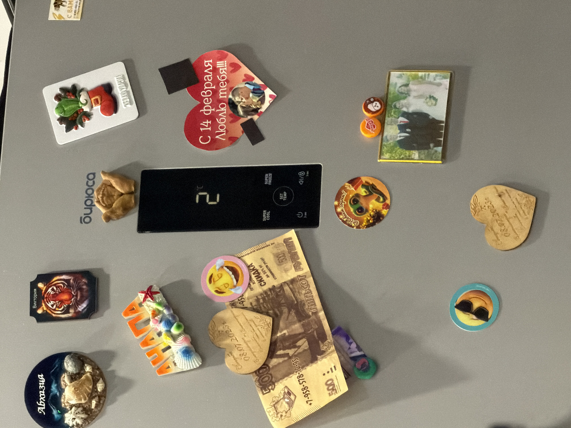 The image is a close-up, eye-level shot of a gray refrigerator door adorned with various magnets and small decorative items. The lighting is even and diffused, suggesting indoor ambient light. No specific city or location information is present in the image itself, nor can it be deduced from the visual content.
On the top left, a round magnet features the word "Абхазия" in stylized white lettering over a dark blue background with images of shells and stones. To its right, a rectangular magnet depicts a tiger's face with the word "Виктория" above it. Further right, a magnet shows a red Christmas stocking with a green bird and berries, with text below that is partially visible. Another magnet to the far right appears to be a small postcard with a picture of a tiger.
Below these, a colorful, three-dimensional magnet spells out "АХАНА" with seashells and a red starfish. A large, black rectangular panel with digital readouts and buttons (displaying "2°C" and text like "SUPER COOL," "SET TEMP," and power icons) is visible on the left side.
A heart-shaped wooden magnet engraved with "08.07.2023" and floral patterns rests on top of a 500-ruble banknote. Adjacent to this is a round magnet with a laughing emoji face. A round, colorful magnet with a frog character and text that may say "С Новым годом!" is positioned below the black panel.
To the right of the banknote, a large red heart-shaped magnet features the text "С 14 февраля Люблю тебя!!!" and a small photograph of a couple kissing. Two small black square magnets are attached to the top right of the heart magnet.
Below the frog magnet, a golden framed photograph shows a wedding party consisting of a couple in the center, a man in a suit and red tie to the left, and a woman in a light pink dress to the right. Two small, round, colorful bottle cap magnets are placed above the framed photo.
On the lower left, a round magnet depicts a yellow smiley face wearing black sunglasses. Another heart-shaped wooden magnet, similar to the one on the banknote, is located in the lower right, also engraved with text that includes "08.07.2023."
The overall impression is that of a personal collection of souvenirs and mementos on a refrigerator door.