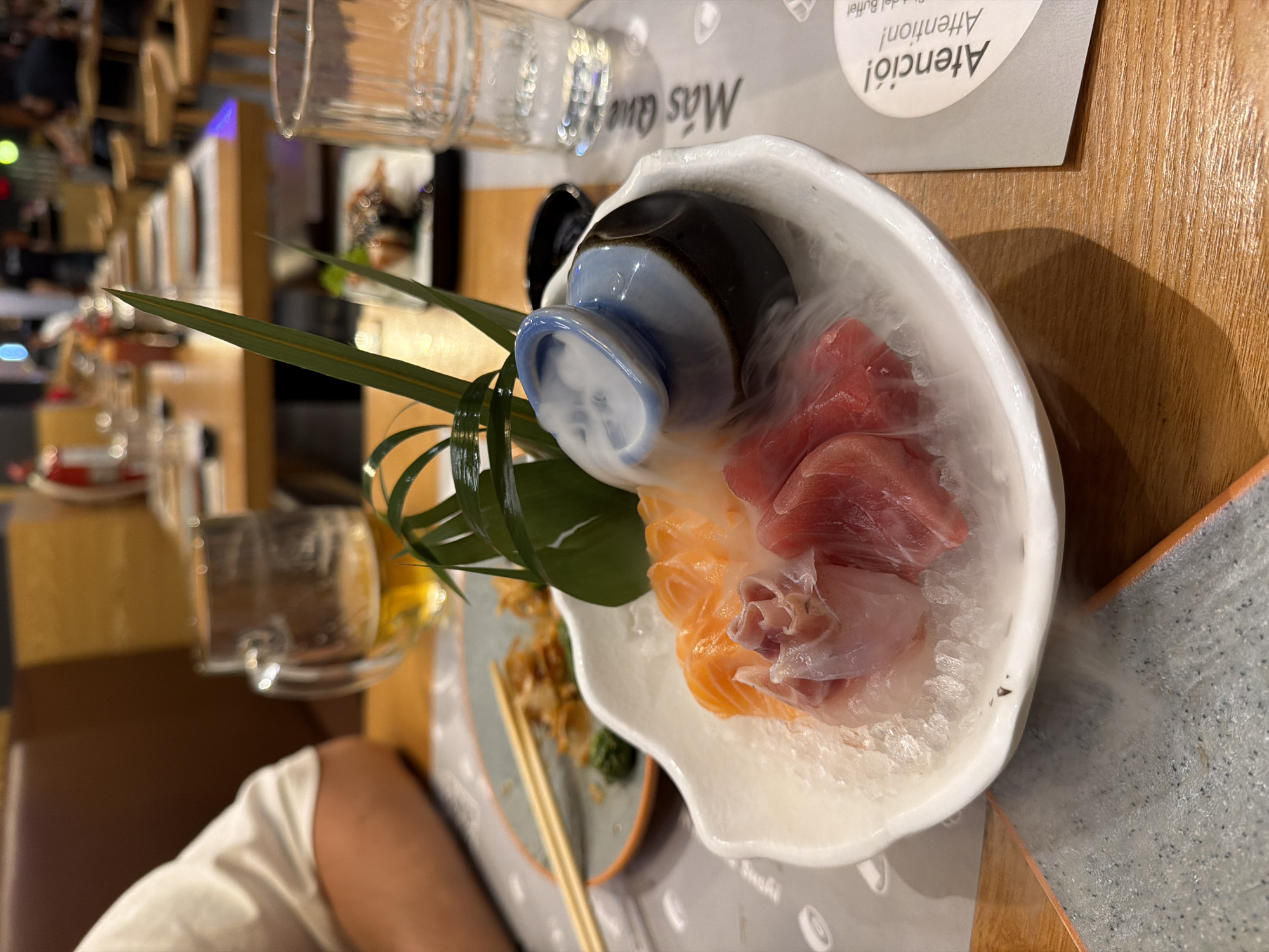 A close-up, eye-level shot captures a plate of sashimi and a small ceramic dish emitting smoke, likely dry ice, on a wooden table at a Japanese restaurant in Barcelona, Spain. The sashimi, consisting of thinly sliced salmon and tuna, rests on a bed of crushed ice in a white, shell-shaped bowl. To the right of the sashimi is a blue and brown ceramic dish with a spout, from which white smoke billows, obscuring the view of the sashimi slightly. In the background, out of focus, are dining tables with place settings, a beer in a glass mug, and patrons, suggesting a lively dining atmosphere. A menu with Spanish text, including "Atención! Attention!", is visible on the right side of the image. The lighting is dim, highlighting the food and the smoky effect.