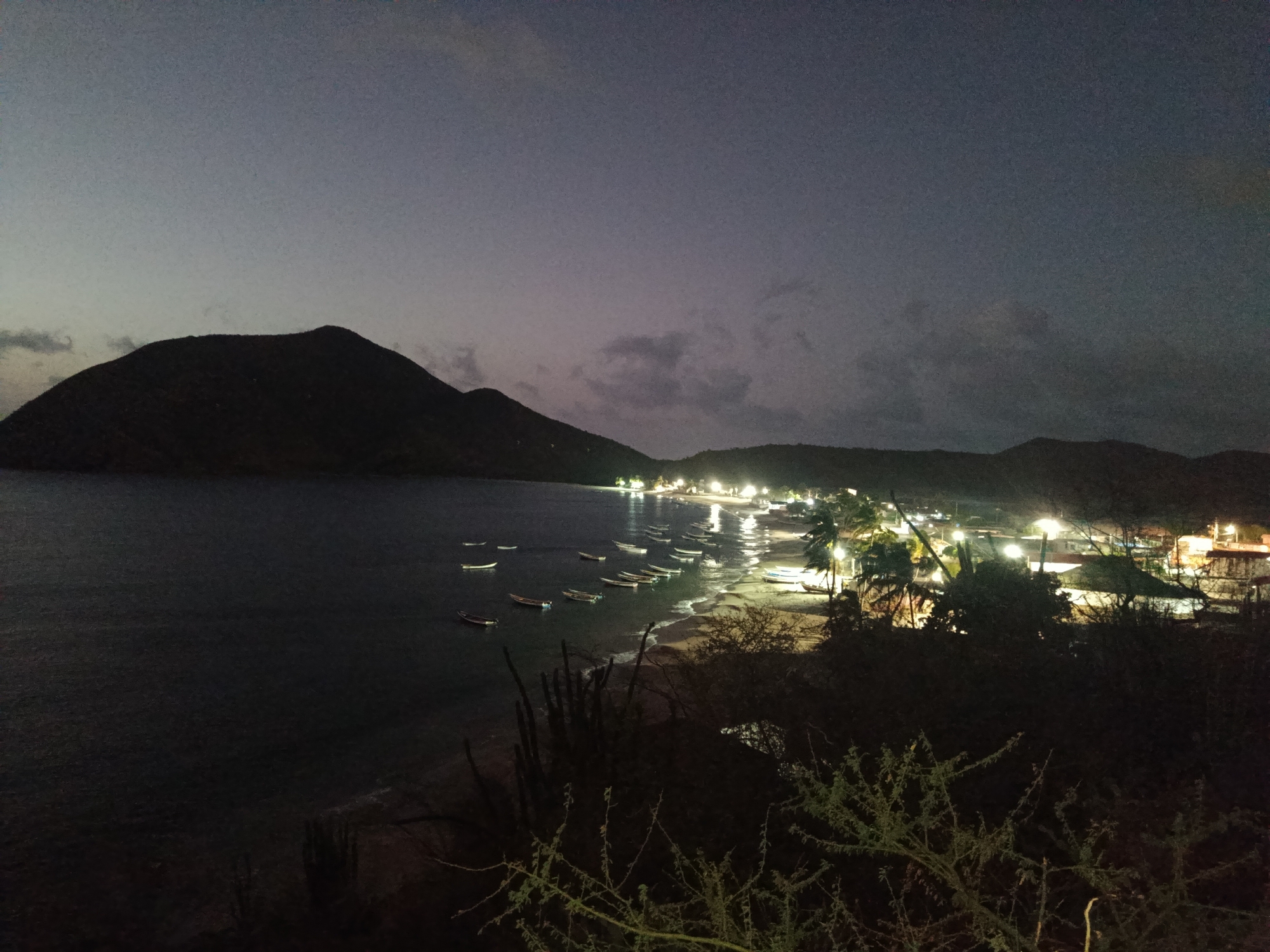 The photograph depicts the coastal city of Juan Griego, Venezuela, at night. In the background, a large dark mountain silhouette is visible. The bay is filled with numerous small boats anchored near the shoreline. A cluster of lights indicates the developed area along the beach. The foreground is darker and obscured by vegetation.