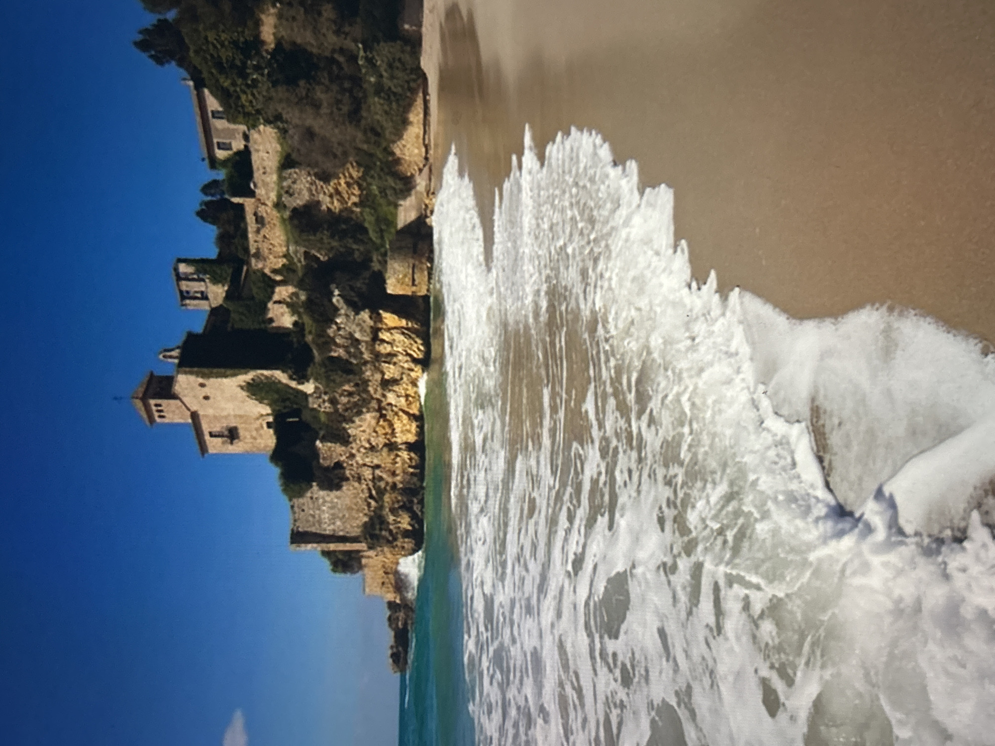 The image presents a vibrant coastal landscape under a clear blue sky. On the left, a collection of historic-looking stone buildings, resembling a fortified village or castle, is perched atop a rugged, rocky cliff. These structures feature traditional architecture with stone walls, visible windows, and tiled roofs, including a prominent square tower that rises above the other buildings. Green vegetation dots the cliff face and surrounds some of the structures.
To the right of the cliff, a sandy beach extends along the shoreline. Ocean waves are actively breaking and receding on the sand, creating dynamic patterns of white foam and ripples on the water's surface. The water closest to the shore is a foamy white, transitioning to a clearer, greenish-blue further out into the sea.
The scene depicts a sunny day with no clouds, emphasizing the natural beauty and historical elements of the location. There are no visible people or discernible human activities taking place. No text is visible in the image.